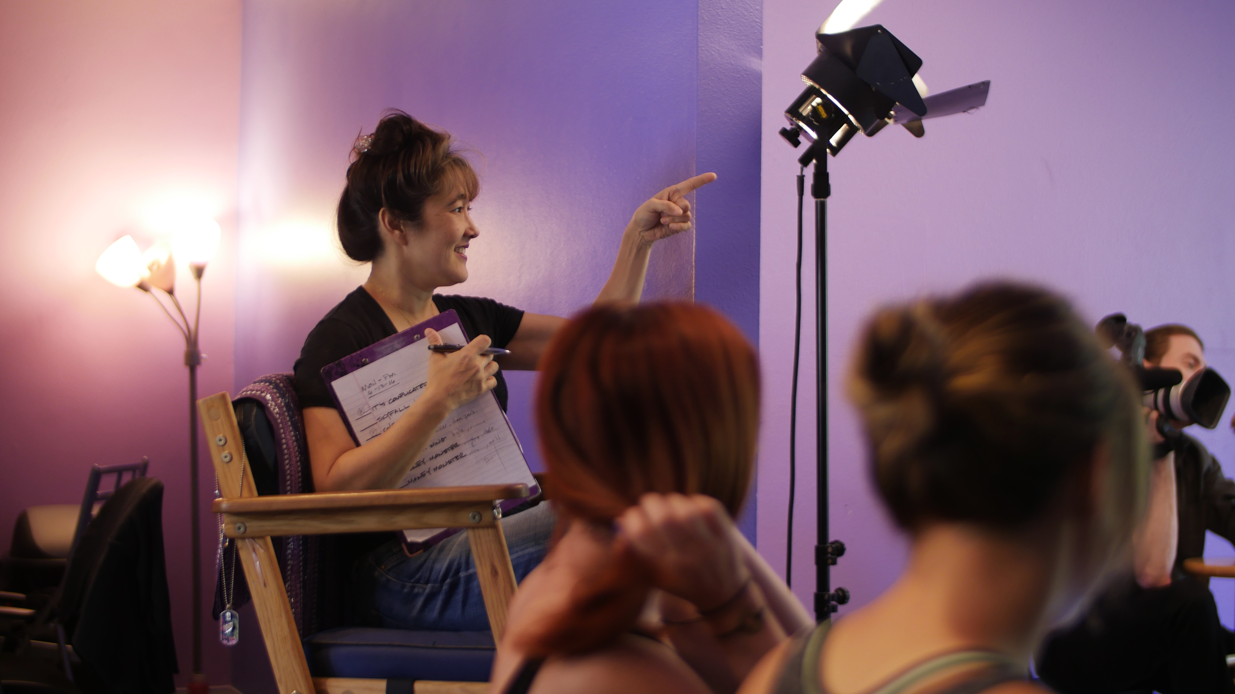 A woman sitting on a director's chair, smiling and pointing at a camera, in a colorful studio setting with purple and pink walls, while holding a script or notebook.