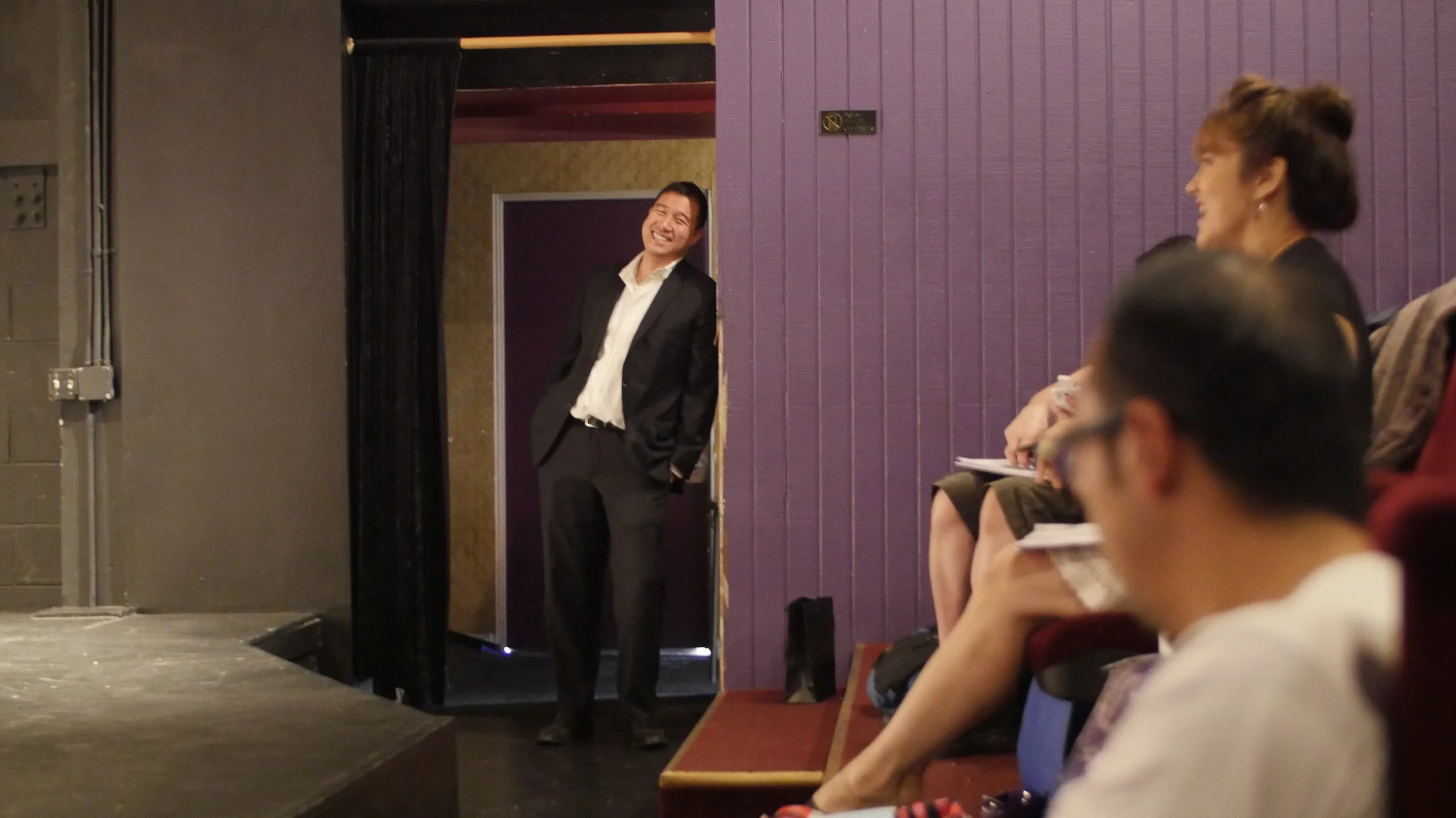 A man in a black suit standing in a doorway, smiling and leaning against the doorframe, while a group of seated women watches and smiles in a theater or conference room.