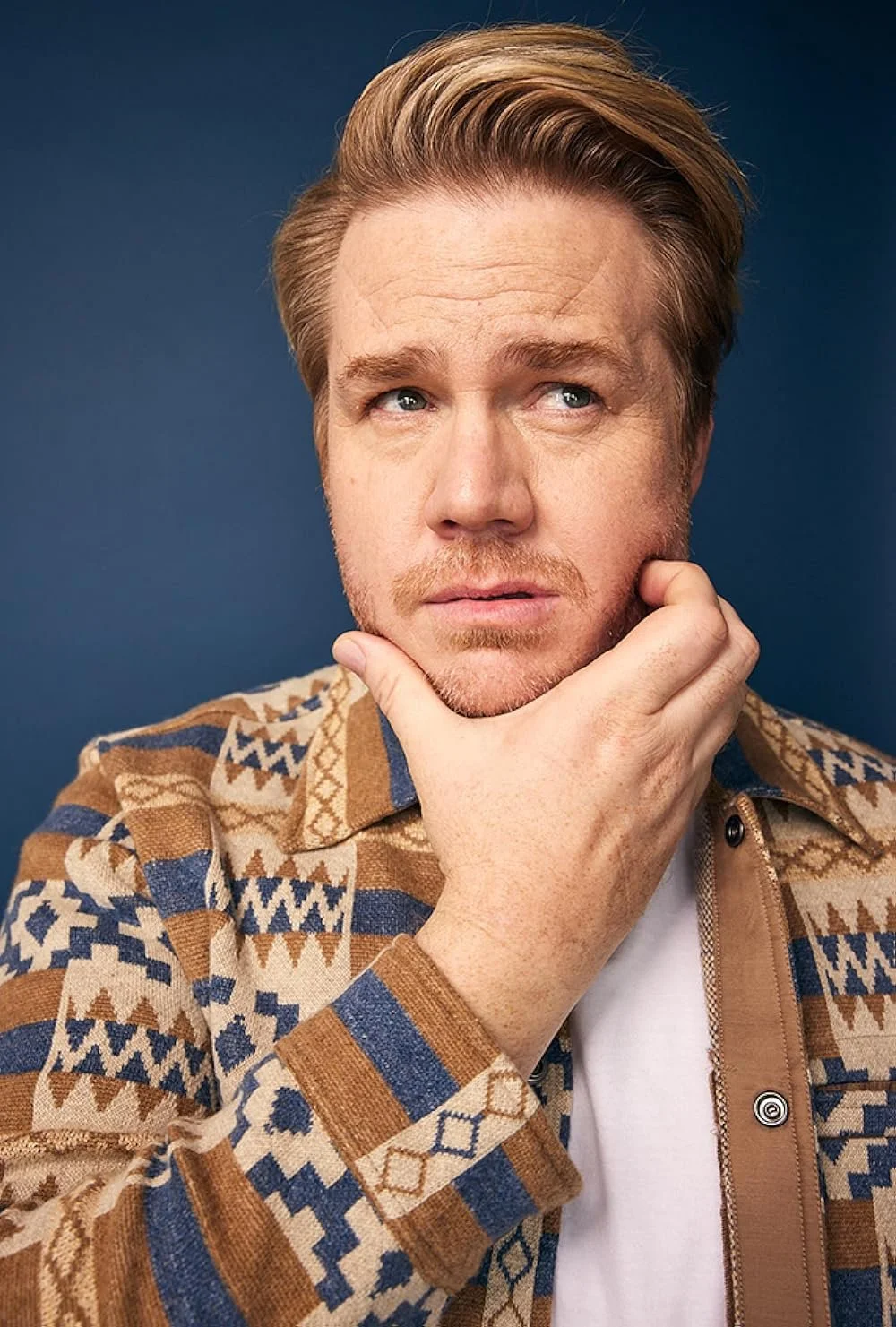 A man with blond hair and stubble on his face, posing with his hand on his chin, wearing a patterned brown, blue, and beige jacket against a blue background.
