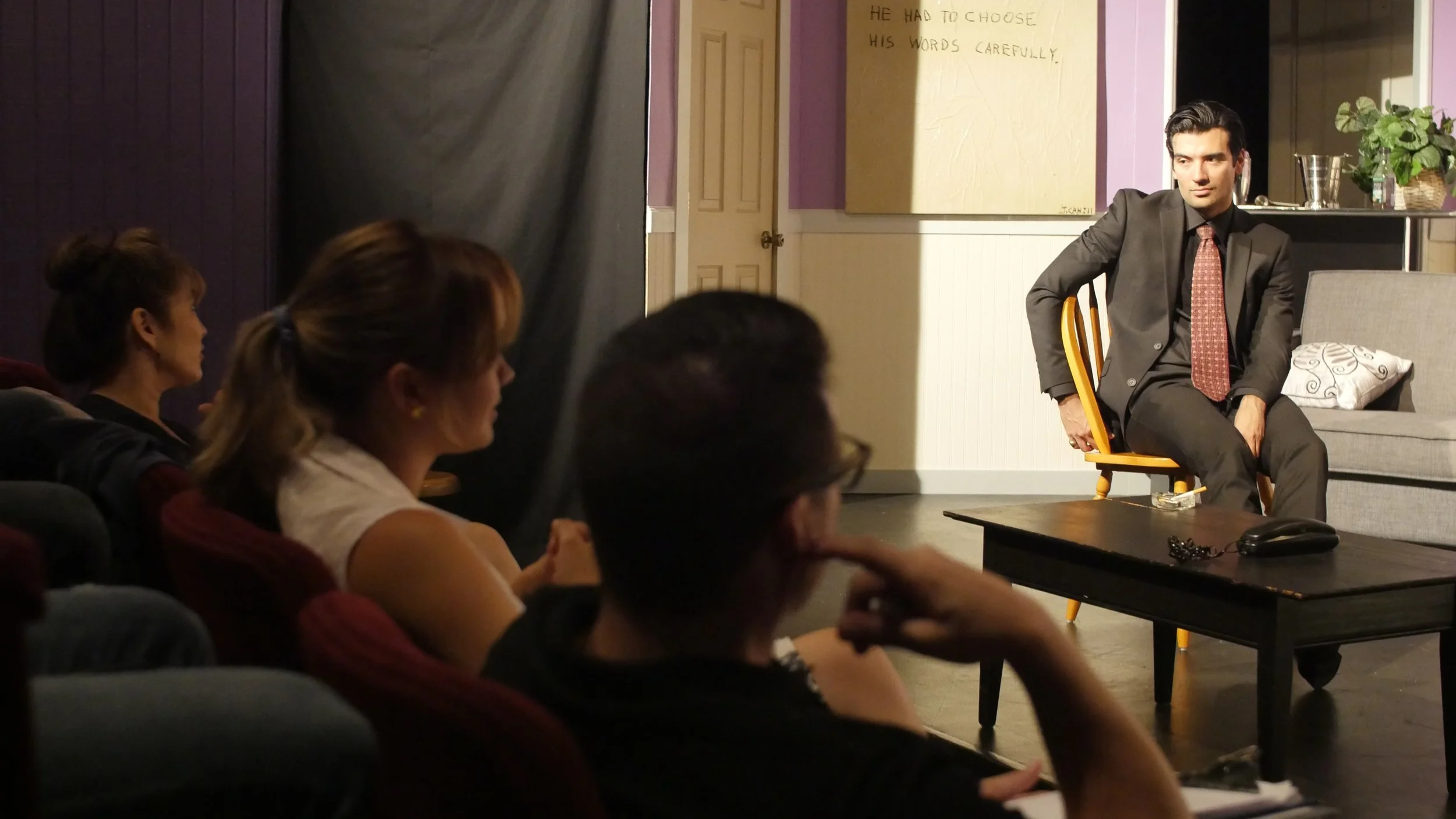 A young man in a suit and tie sitting on a wooden chair and talking to an audience in a small theater or classroom setting, with four women seated and listening attentively.