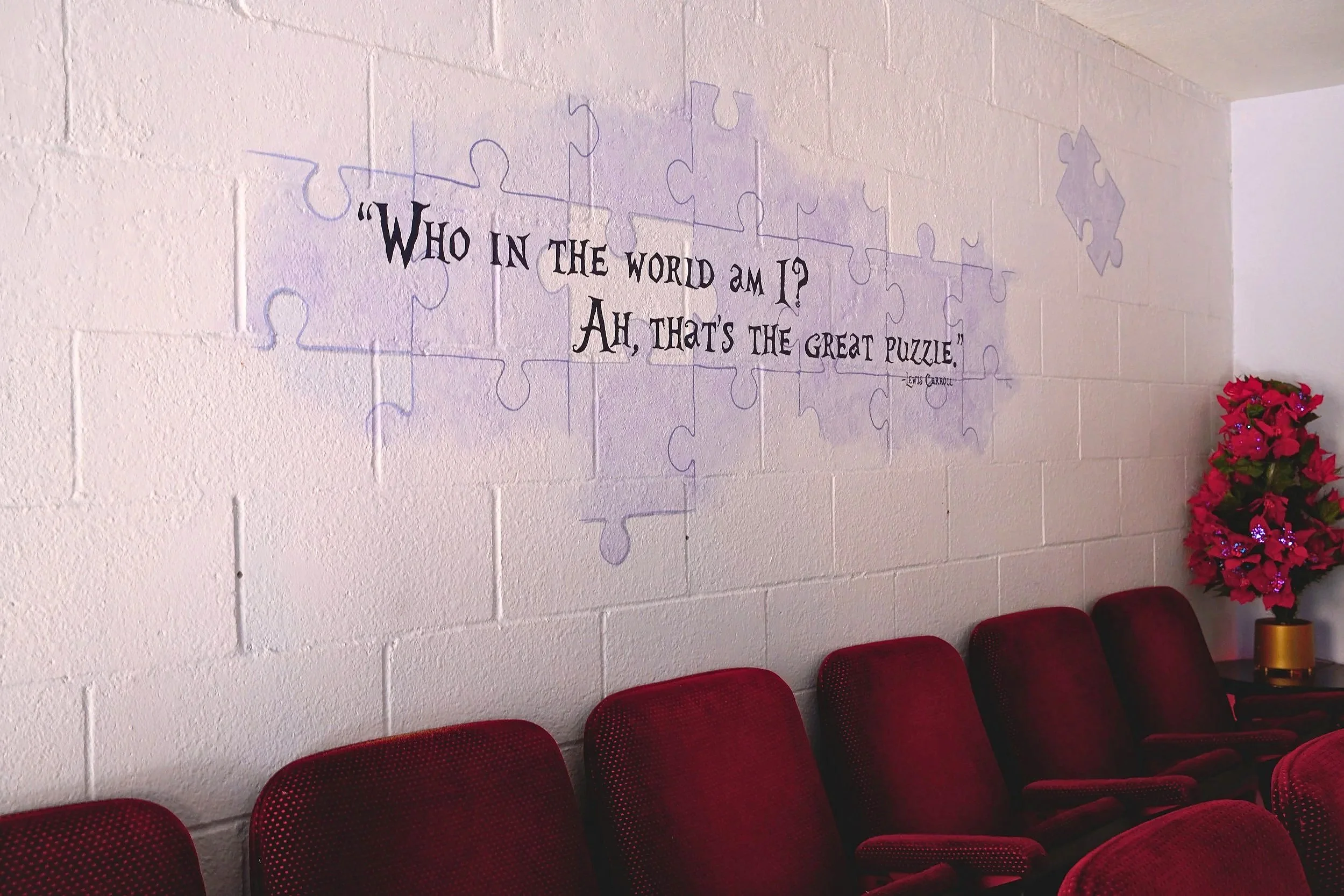A wall with a quote by Lewis Carroll written on it, surrounded by puzzle piece outlines. The quote reads 'Who in the world am I? Ah, that's the great puzzle.' Below the quote, there are red cushioned chairs lined up and a pink poinsettia plant on a table.