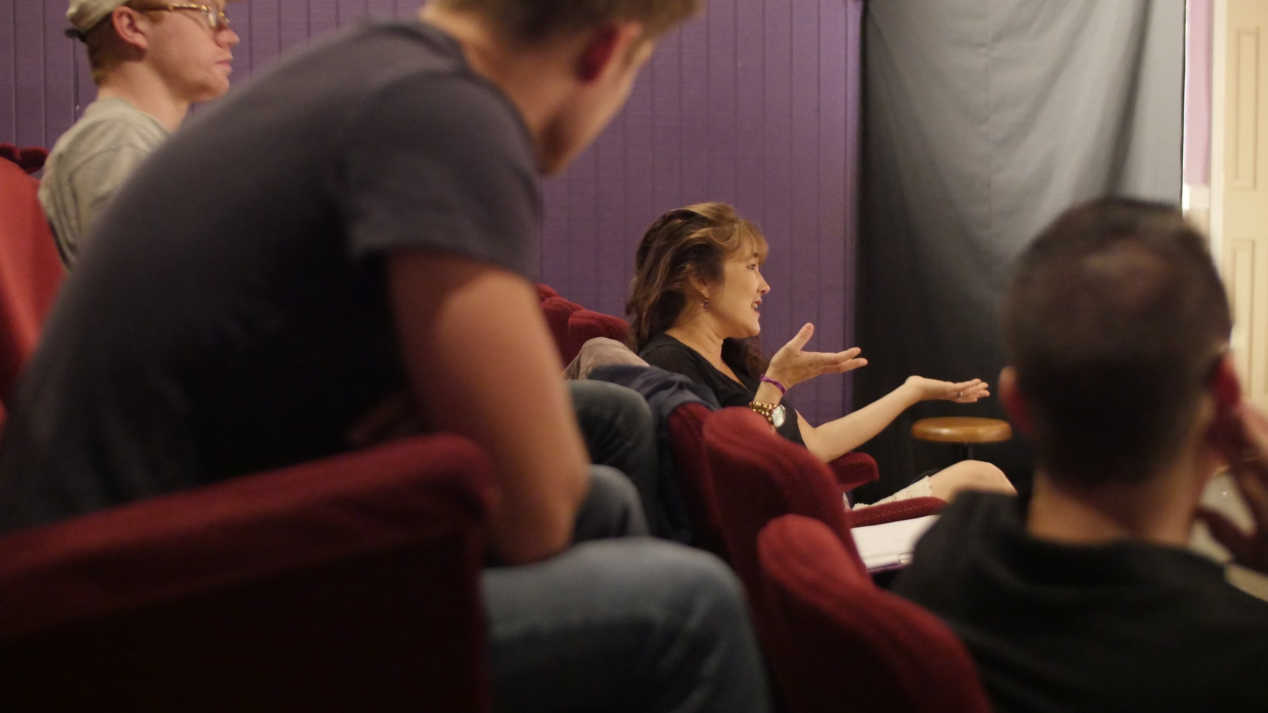 A woman speaking in a discussion, surrounded by several seated people in a dimly lit room with purple walls.
