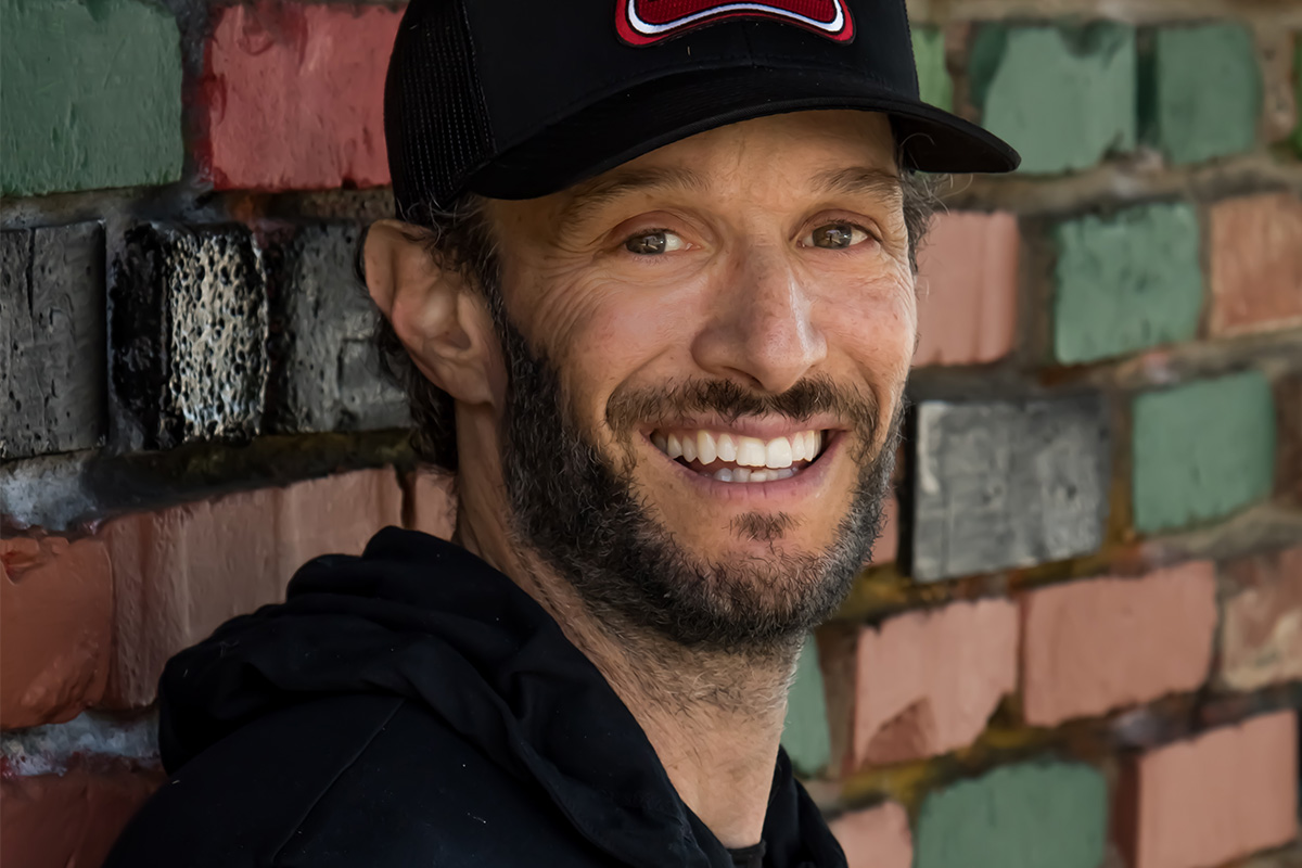 A smiling man with a beard leaning against a colorful brick wall, wearing a black cap and black hoodie.