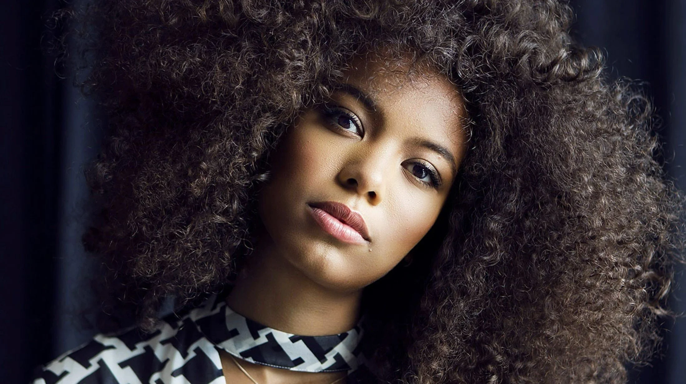Close-up portrait of a woman with voluminous, curly hair, light skin, and makeup, wearing a black and white patterned top.