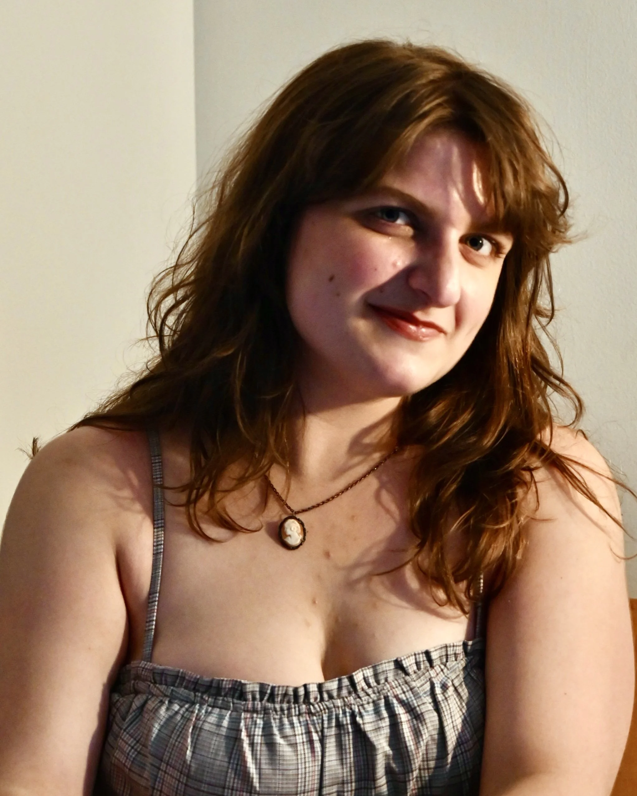 A woman with wavy red hair, wearing a checkered sleeveless top and a cameo necklace, sitting indoors and smiling at the camera.