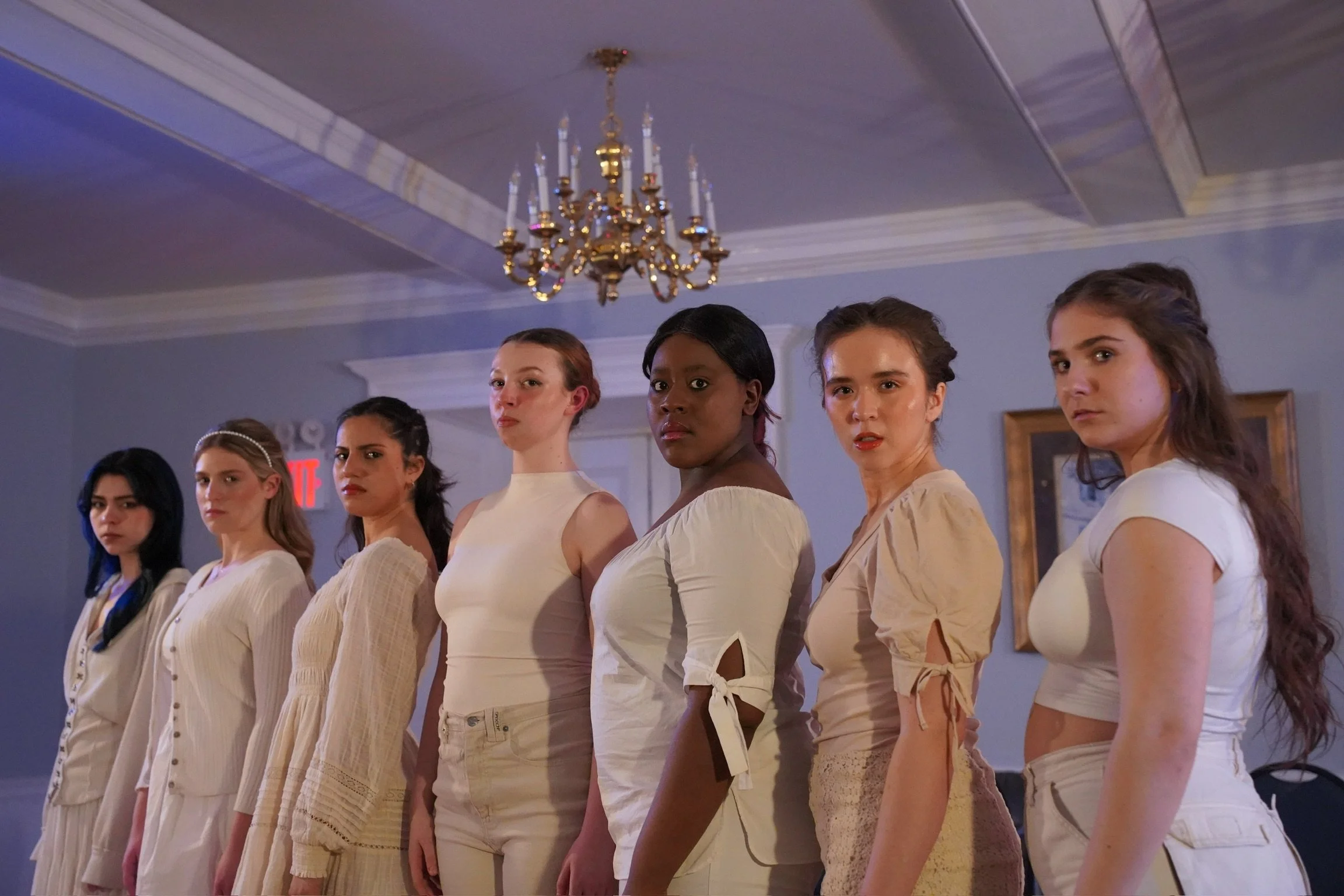 Seven women standing in a line indoors, dressed in light-colored clothing, with a chandelier hanging from the ceiling behind them.