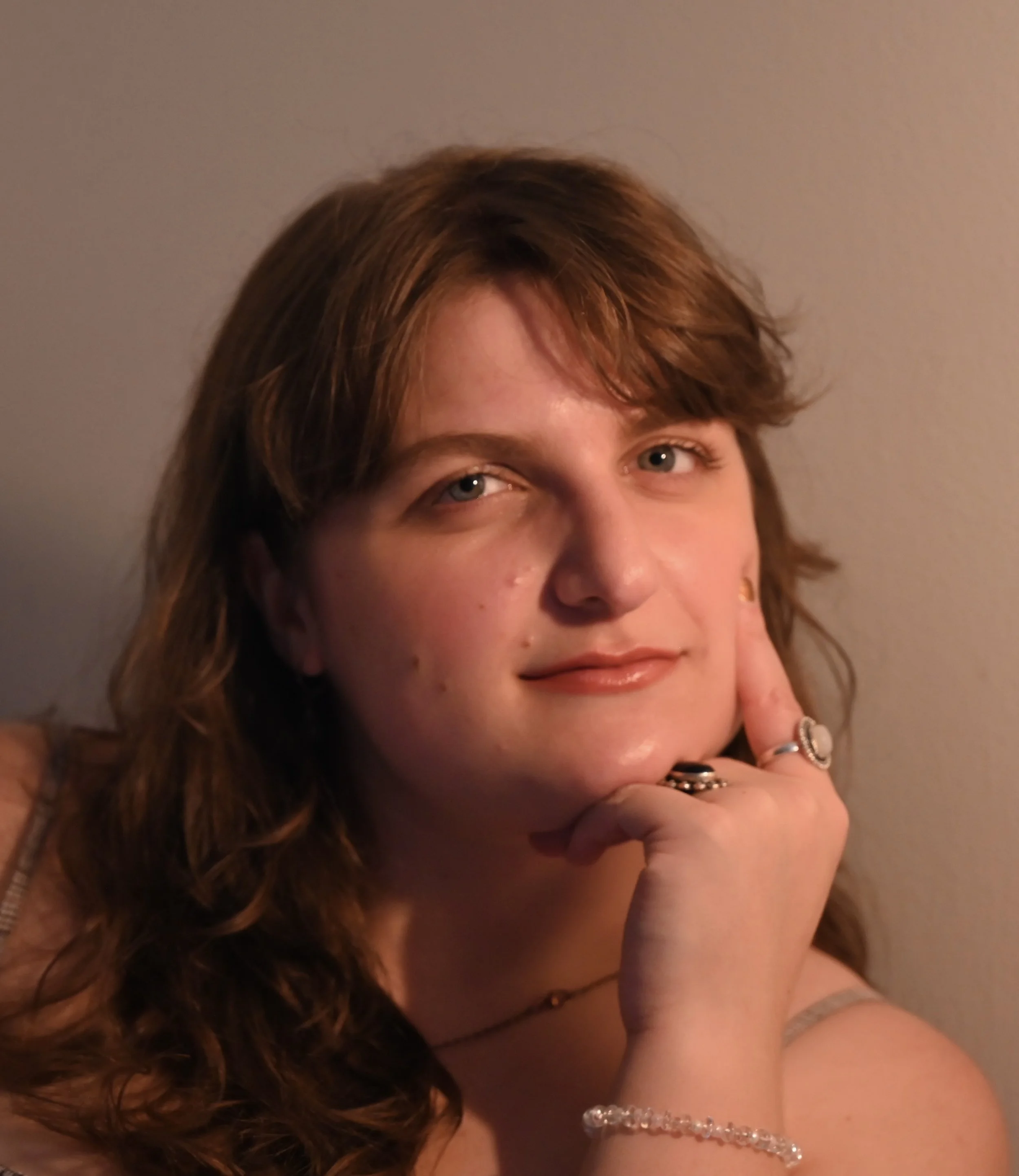 A woman with reddish-brown hair resting her chin on her hand, with a gentle smile and jewelry, looking at the camera.