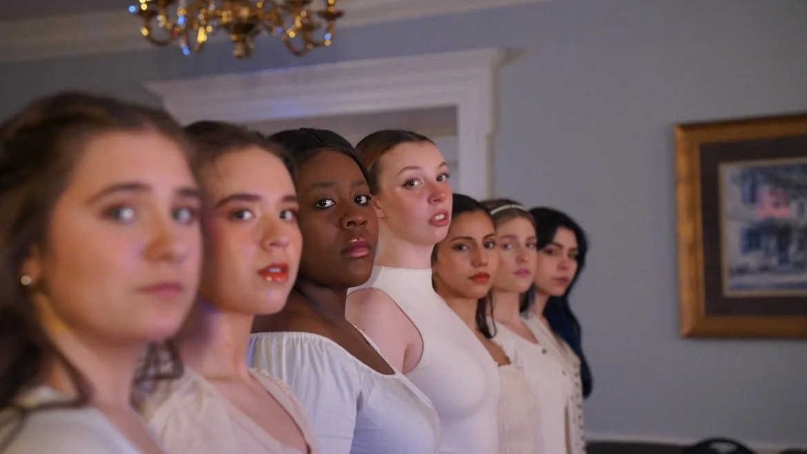 Seven women standing in a line indoors, looking towards the camera with serious expressions.