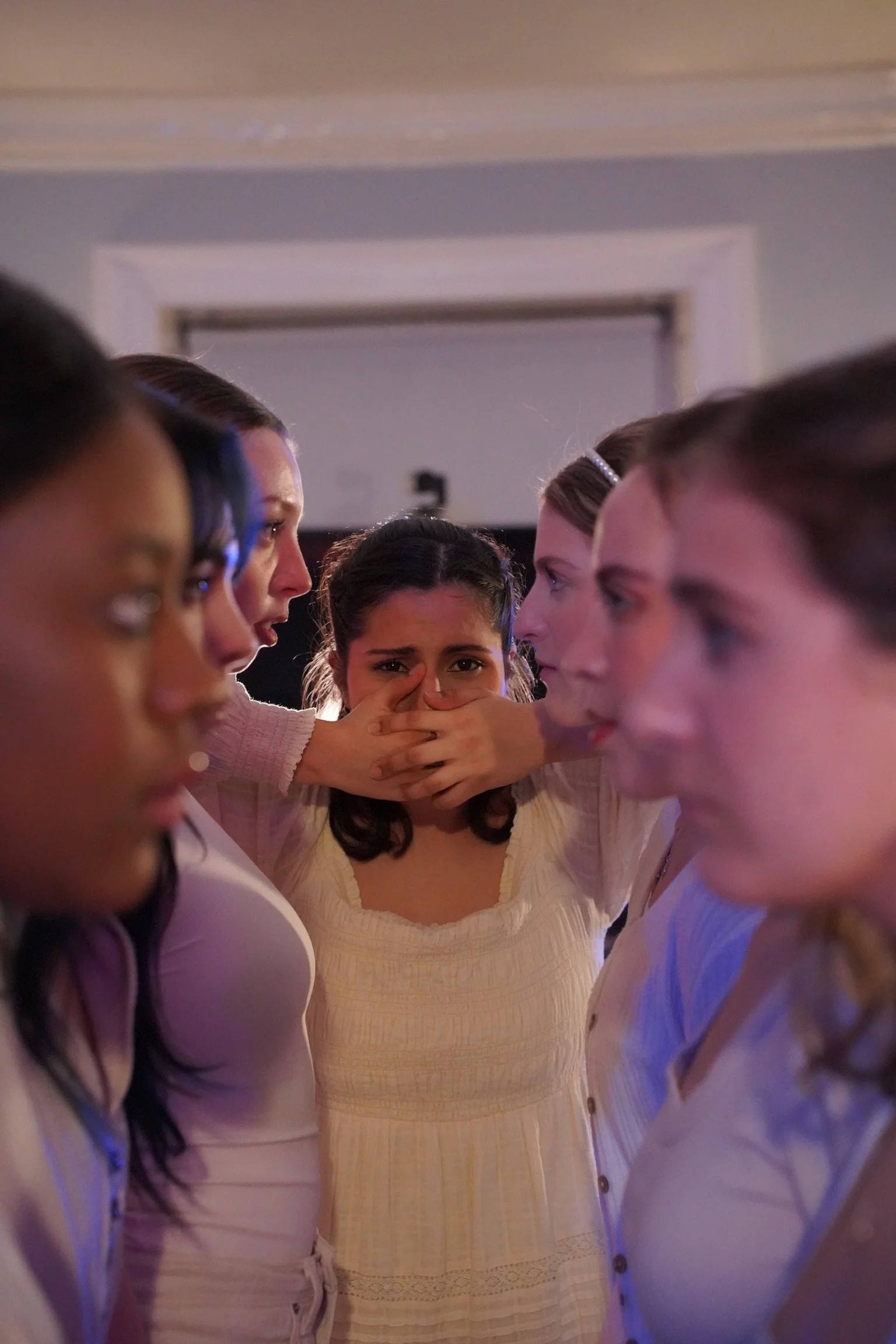 Group of young women standing close together with serious and distressed expressions, one in the center covering her mouth with her hands.