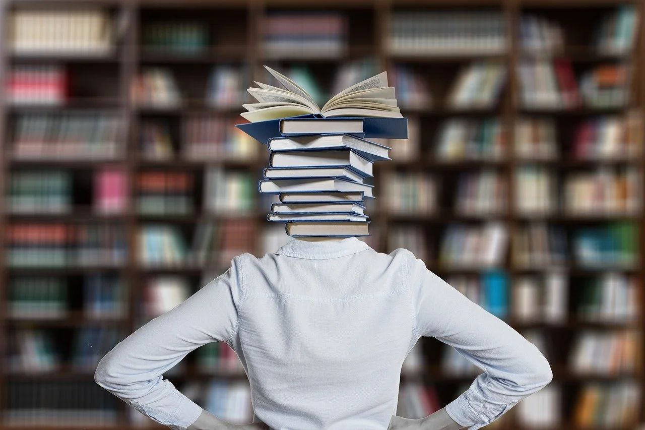 Person with a stack of books as their head, standing in front of a bookshelf.