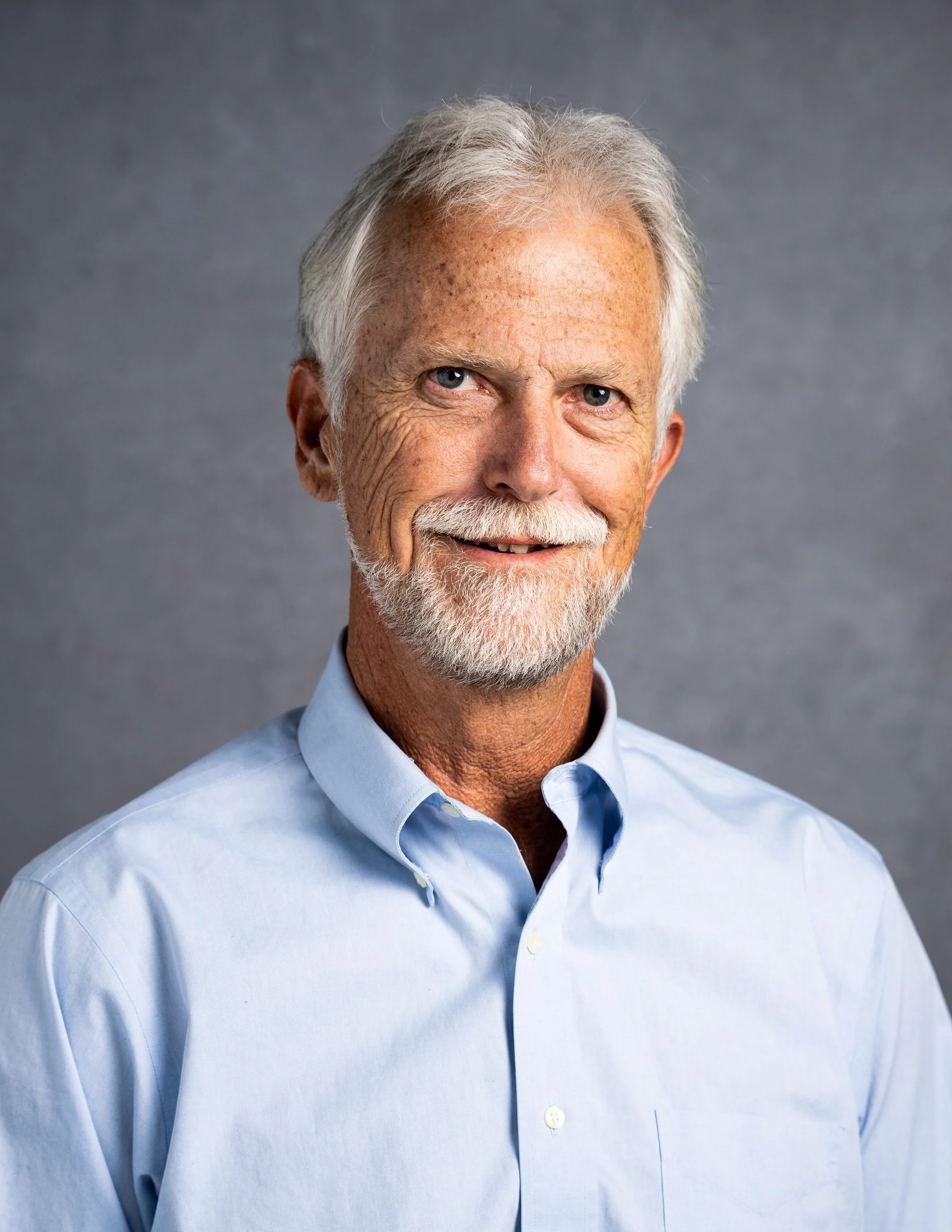 Headshot of an elderly man with white hair and a beard, smiling, wearing a light blue button-down shirt, against a gray background.