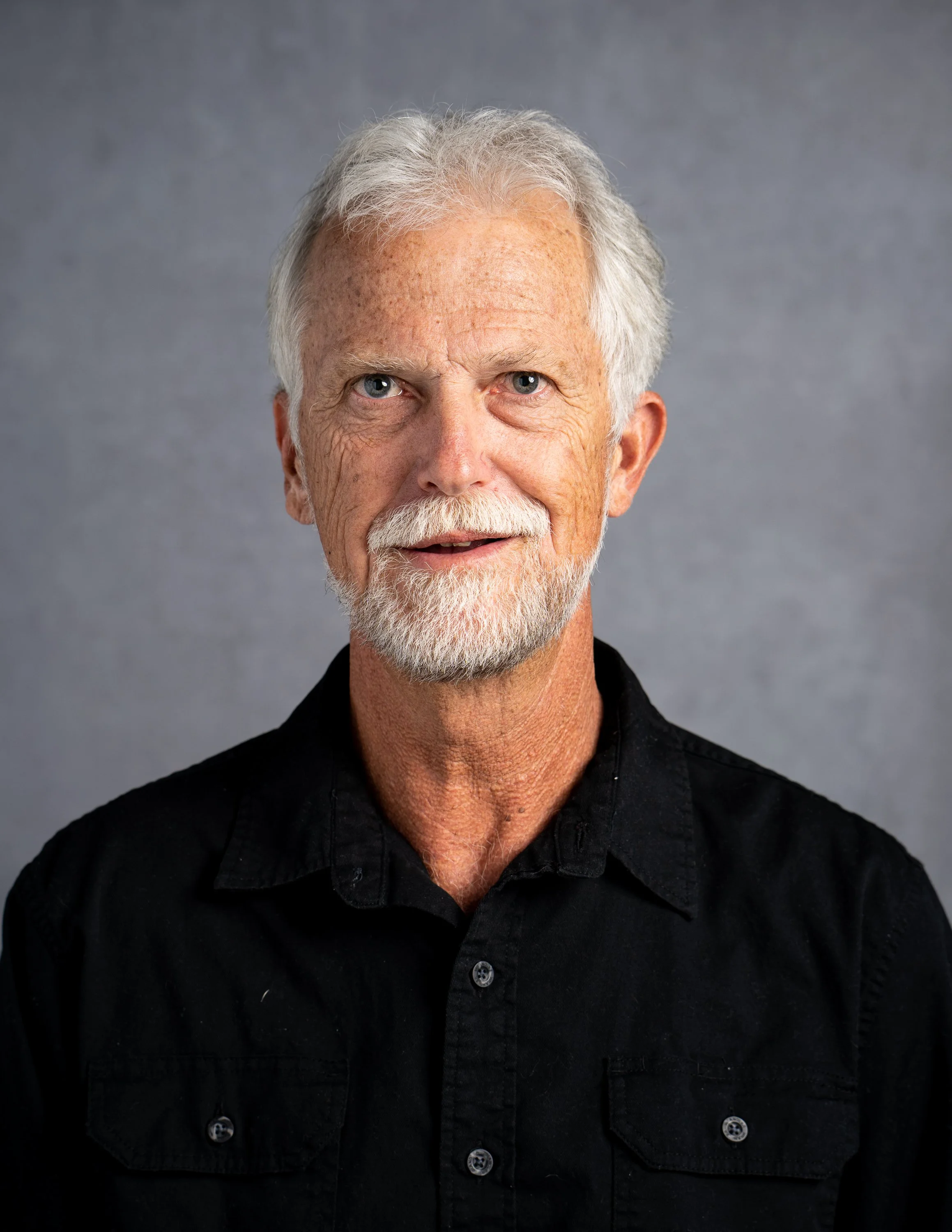 Portrait of an elderly man with white hair, beard, and mustache, wearing a black collared shirt, standing against a gray background.