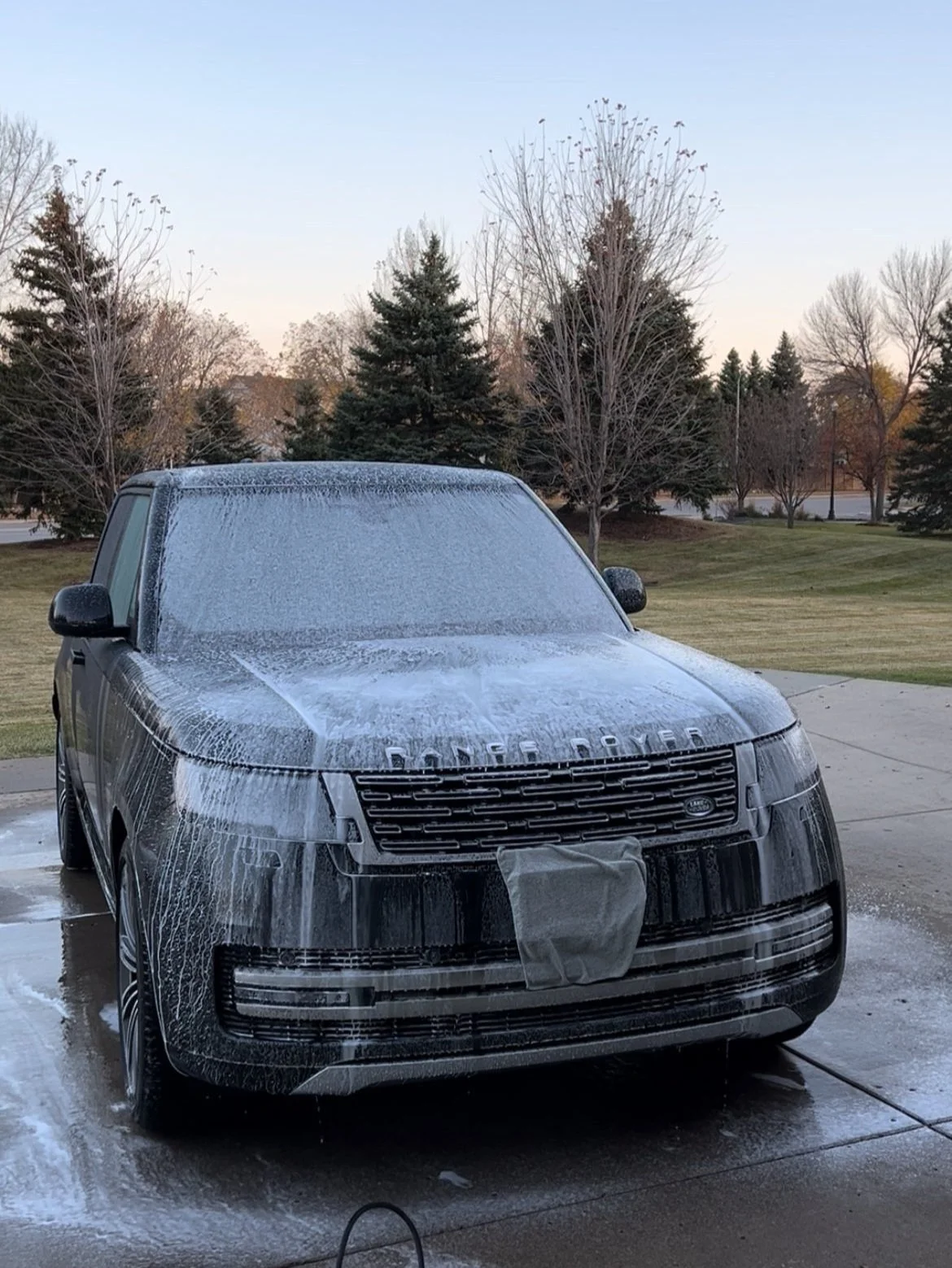 Black Range Rover SUV covered in white foam during car wash outside on a driveway with bare trees and a grassy area in the background.