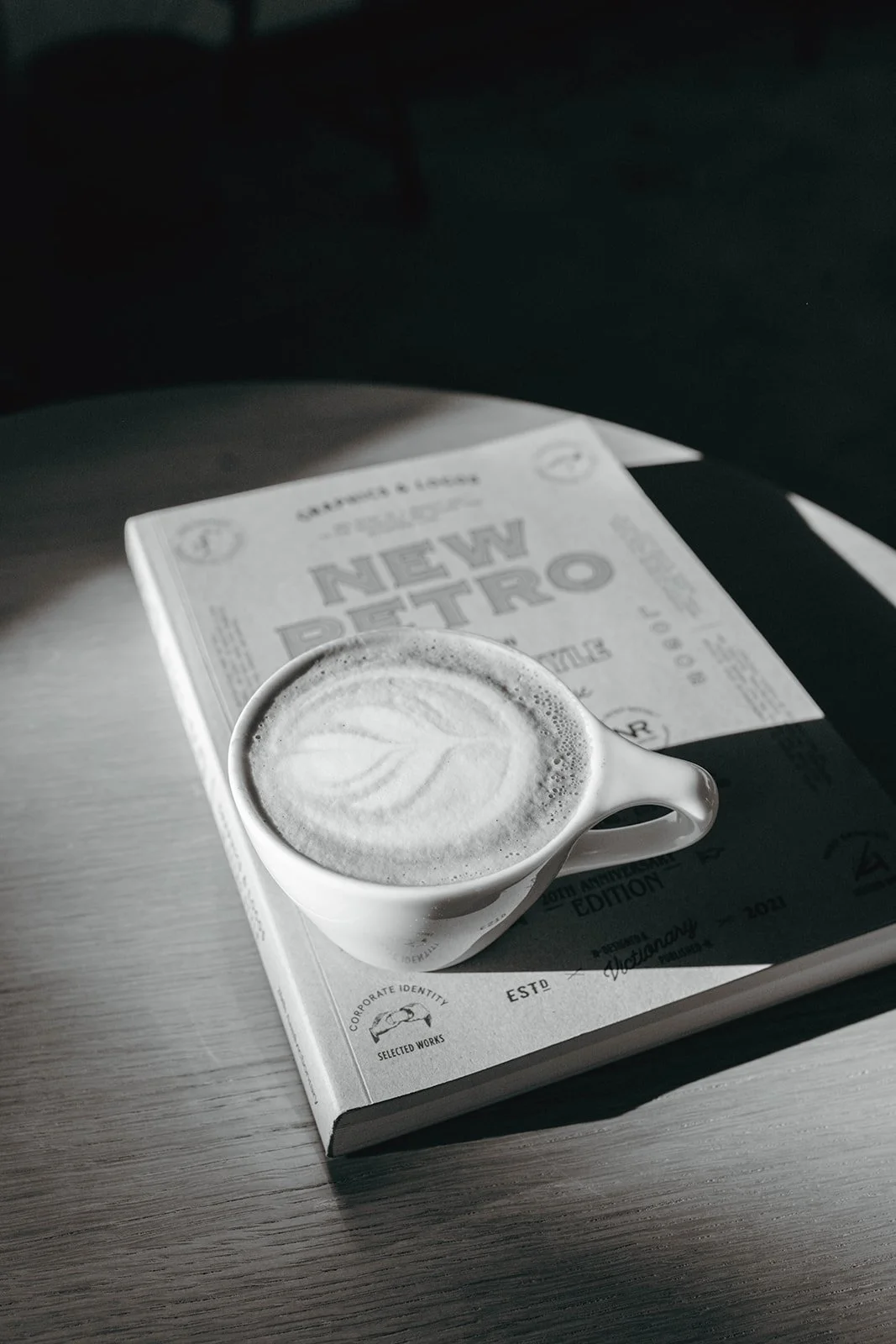 A cup of latte with latte art on a book titled 'New Petro' on a wooden table, with sunlight casting shadows