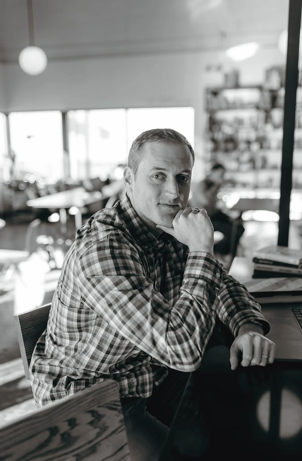 A man wearing a plaid shirt sitting at a table in a cafe, resting his chin on his hand, looking towards the camera, with shelves and tables in the background.
