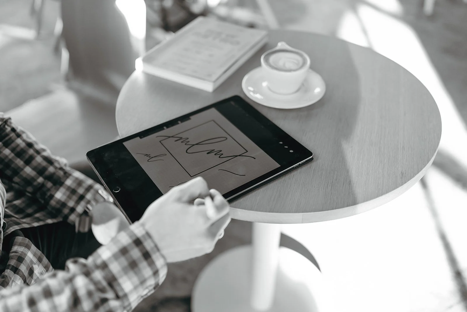 A man in a checkered shirt sitting in a cafe, using a stylus on a tablet, with a coffee and a book titled 'New Retro' on the table.