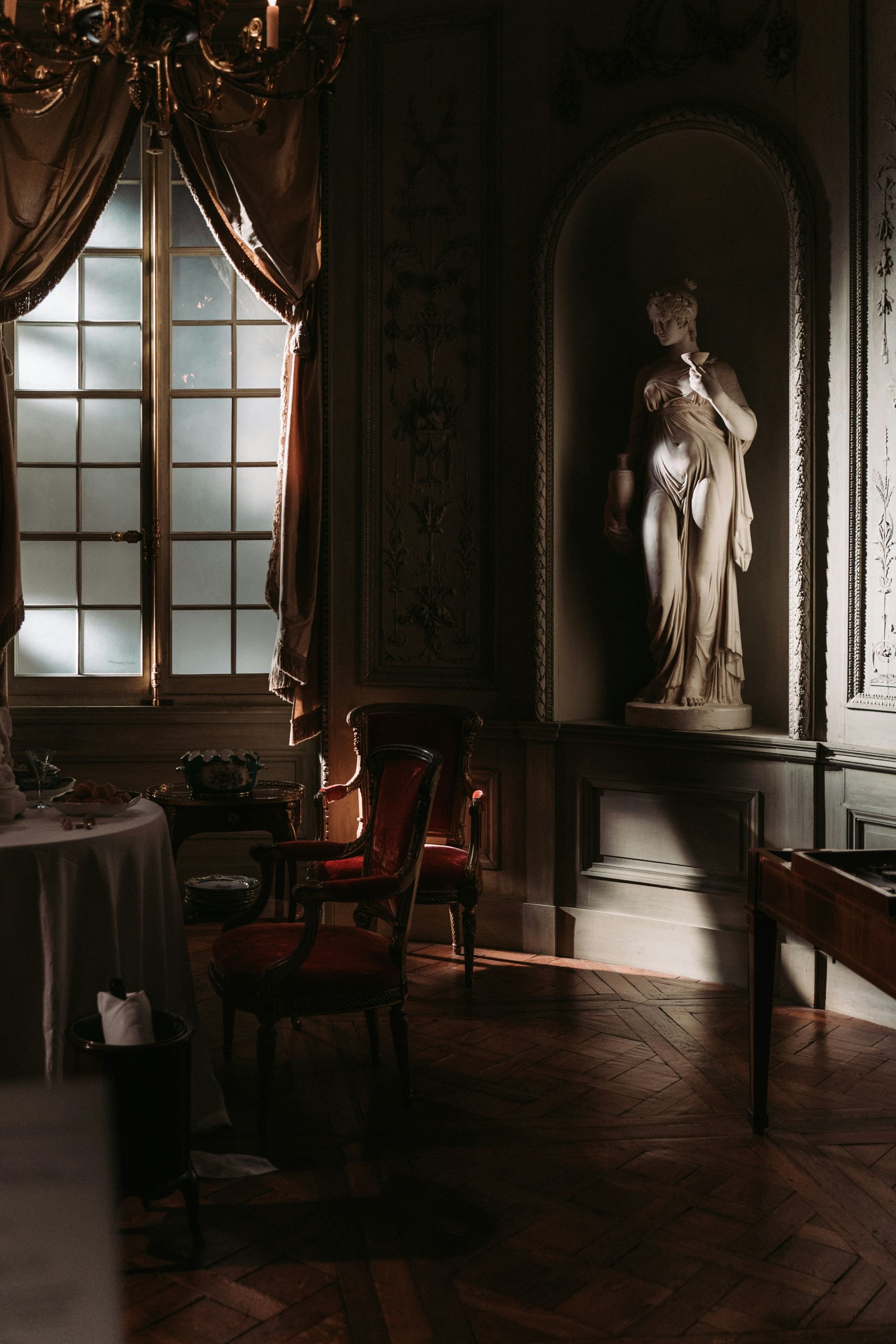 a dimly lit classical interior room with a window, vintage chairs, a table, and a sculpture of a woman in a niche.