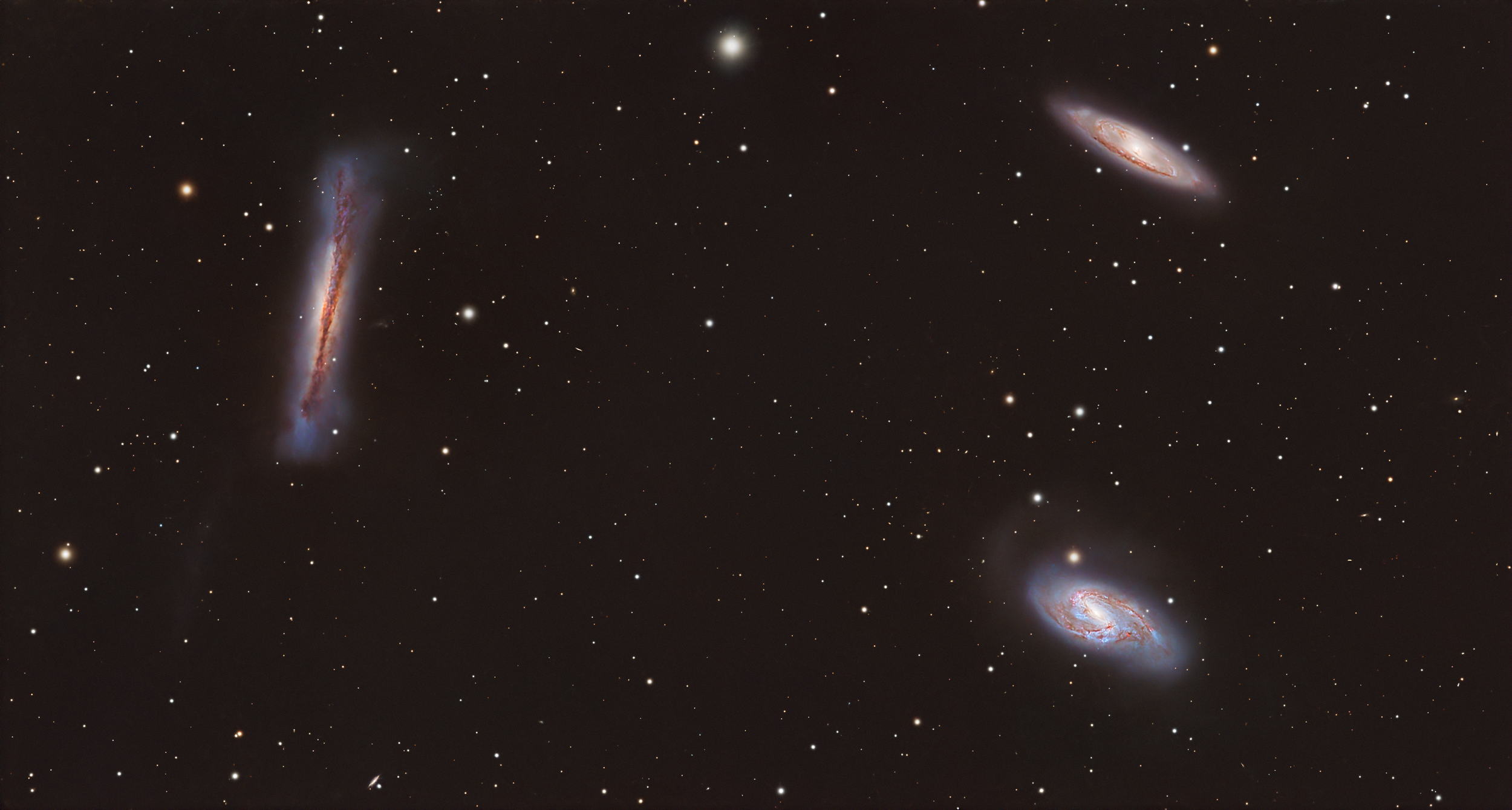 A wide view of space showing several galaxies, including a large, elongated galaxy on the left and two spiral galaxies on the right, with numerous stars scattered across the image.