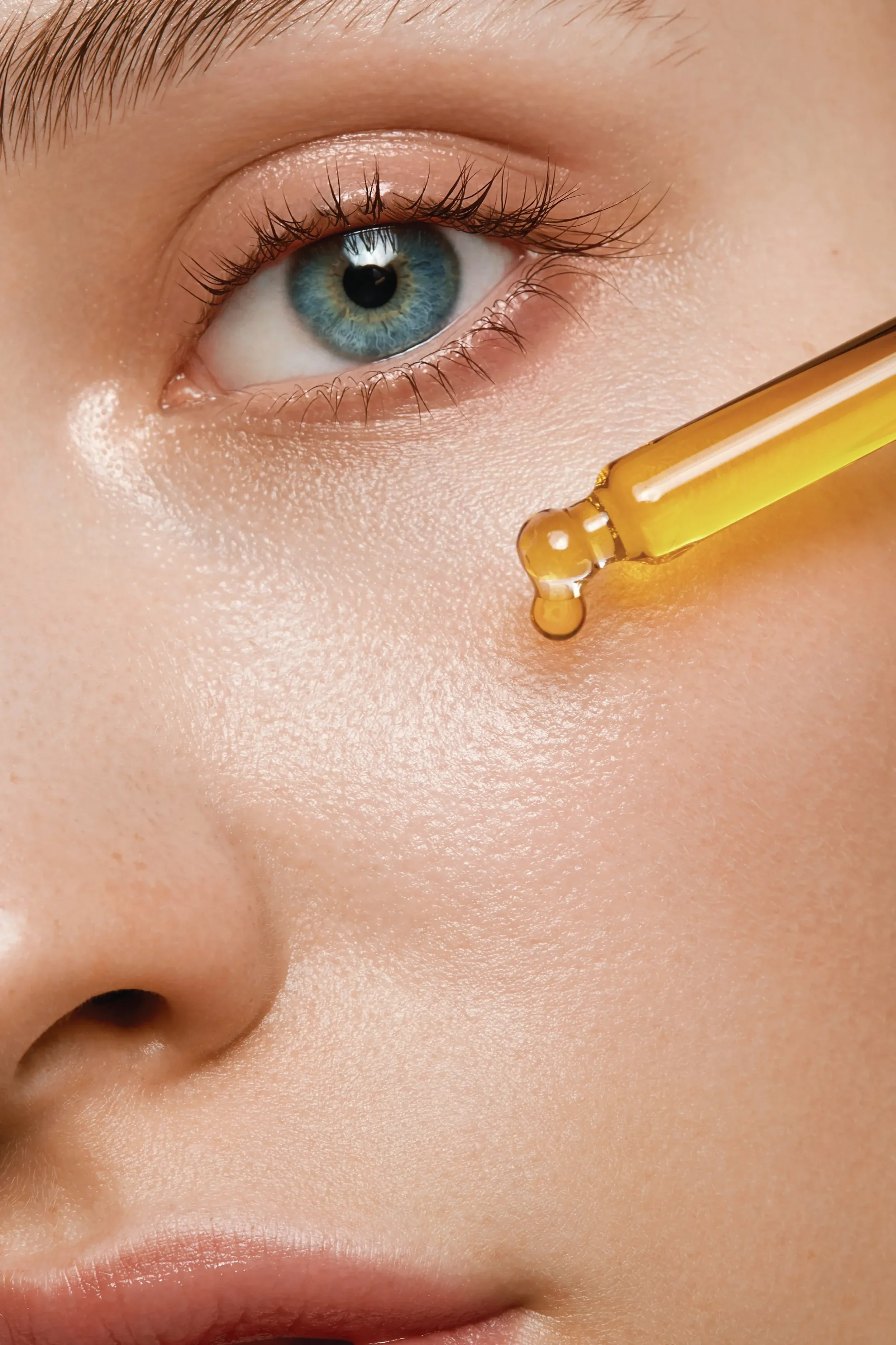 Close-up of a person with blue eyes and fair skin, applying facial serum with a dropper.