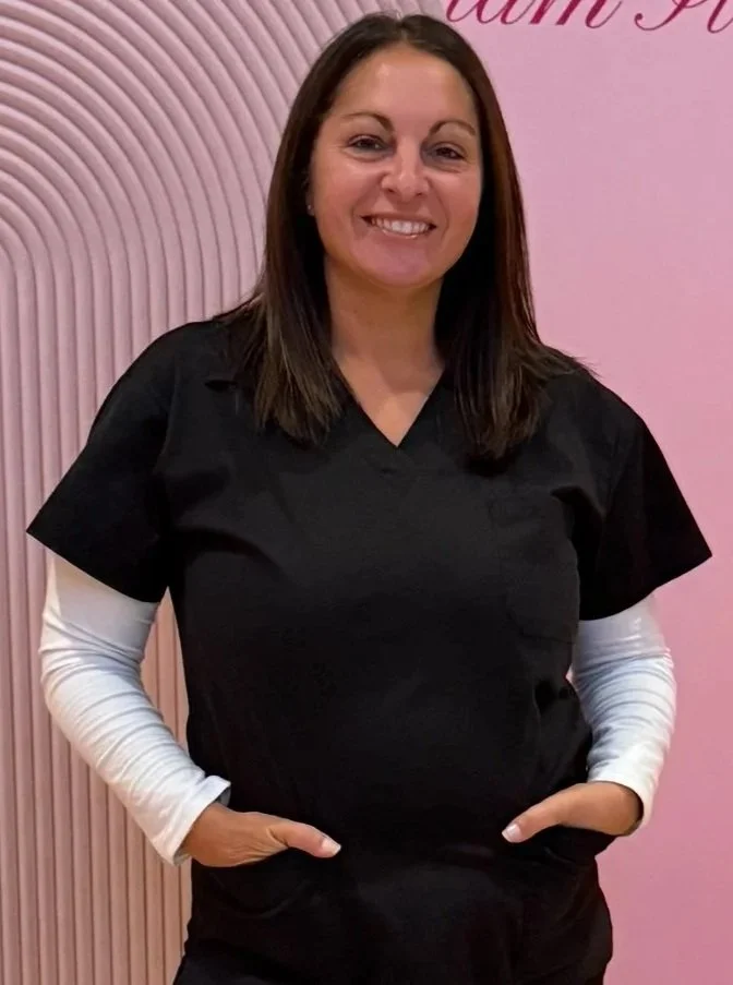 A woman with long brown hair smiling, wearing black scrubs with white long-sleeve undershirt, standing in front of a pink background with curved striped pattern.
