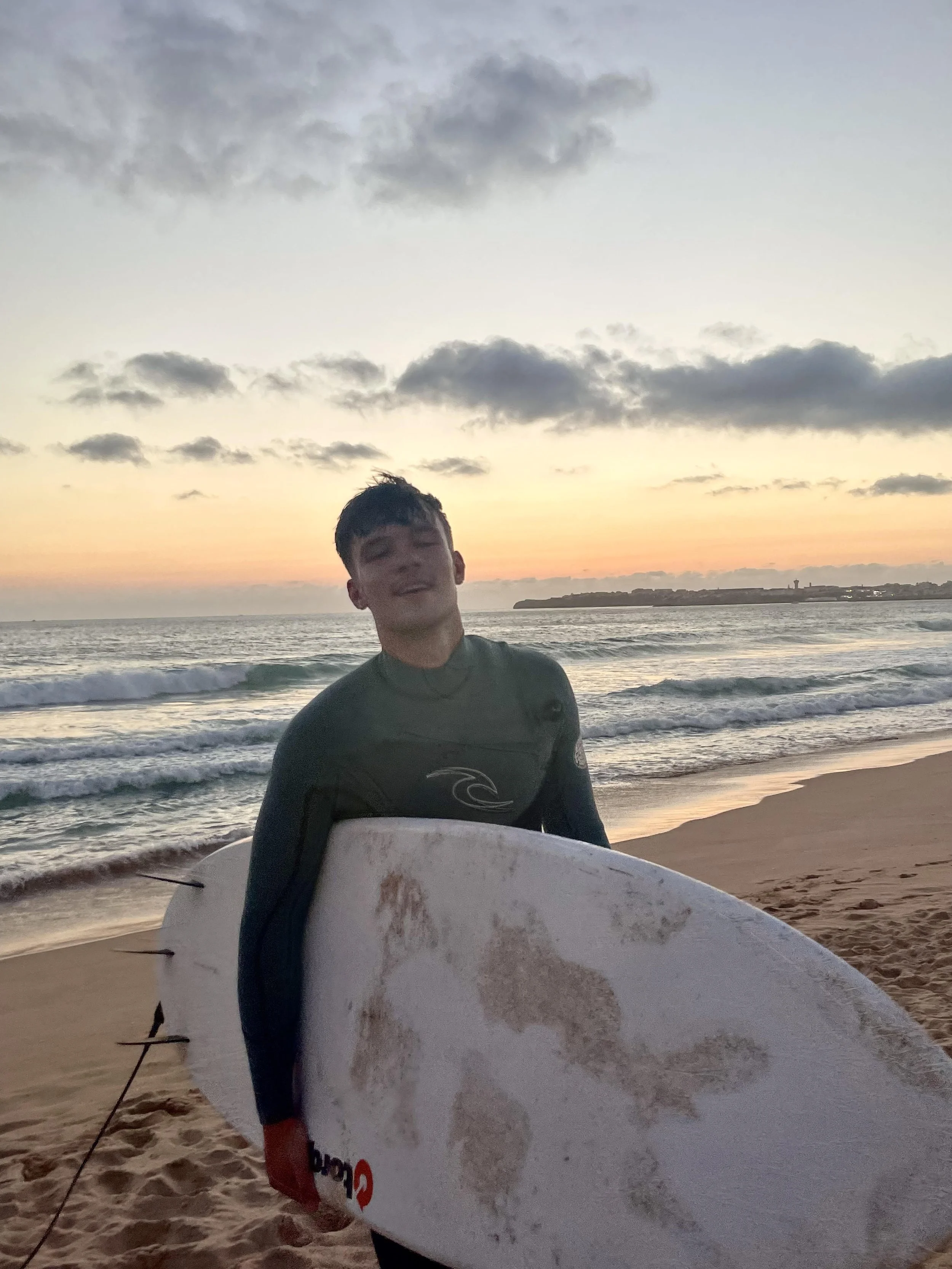 A young person in a wetsuit holding a surfboard on a beach during sunset.