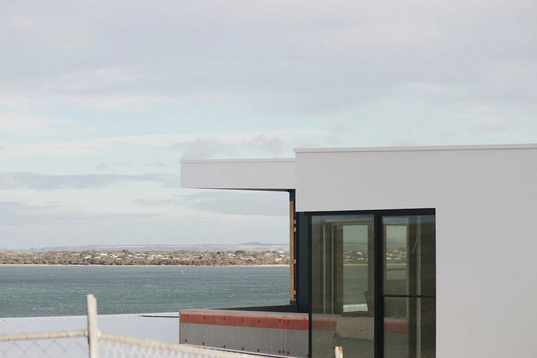 Partial view of a modern white building with large windows overlooking water and land with a cloudy sky.