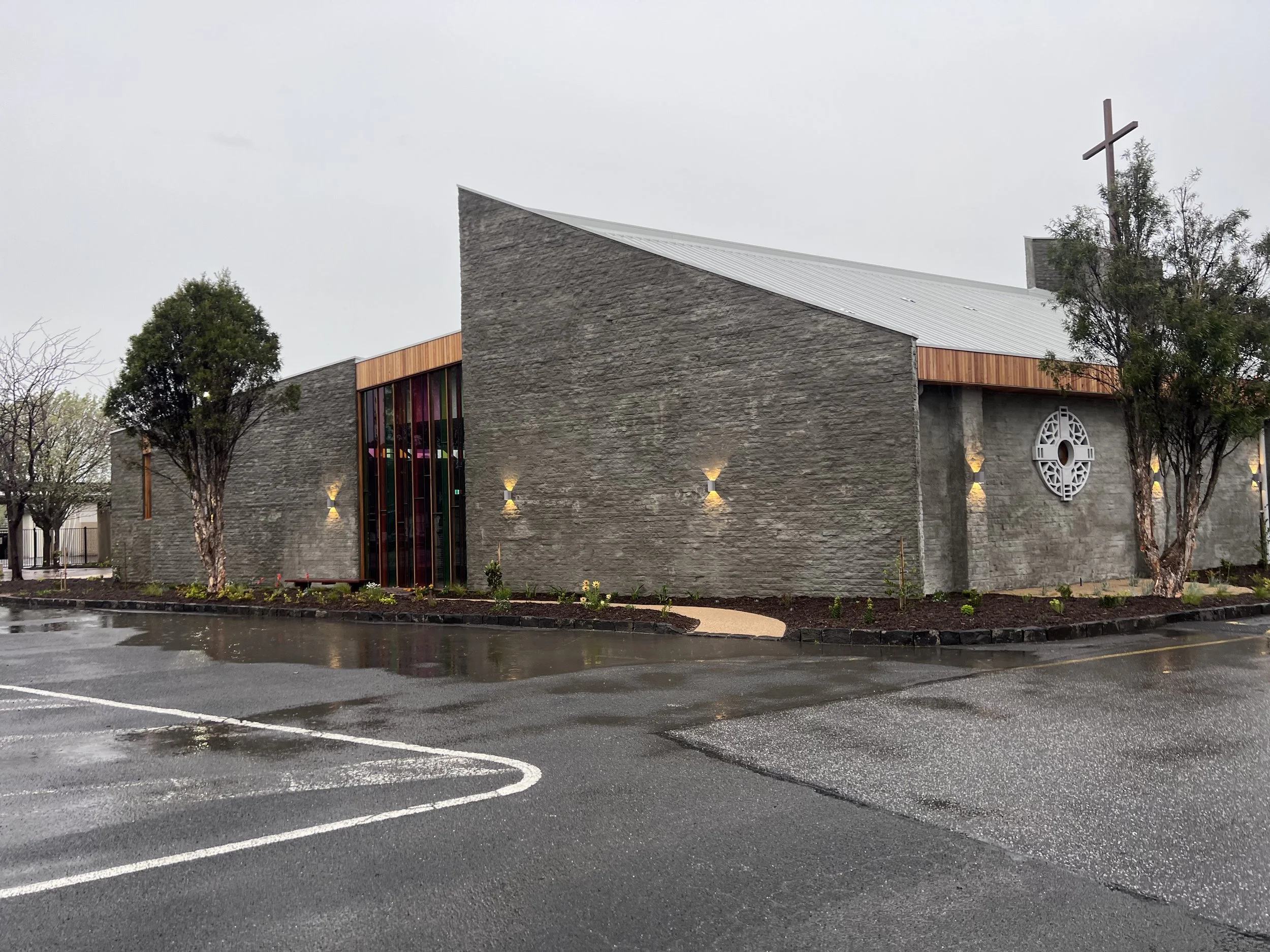 A church building with stone walls, colorful stained glass windows, and a wooden cross on the roof, surrounded by trees and empty parking lot on a rainy day.