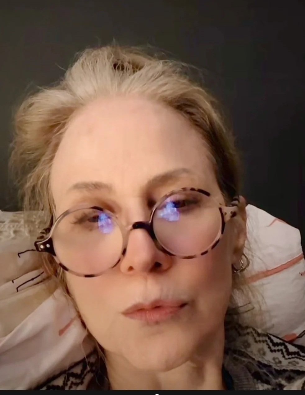 A woman with light brown hair lying on a bed, wearing large round glasses with reflections on the lenses, and a patterned shirt.