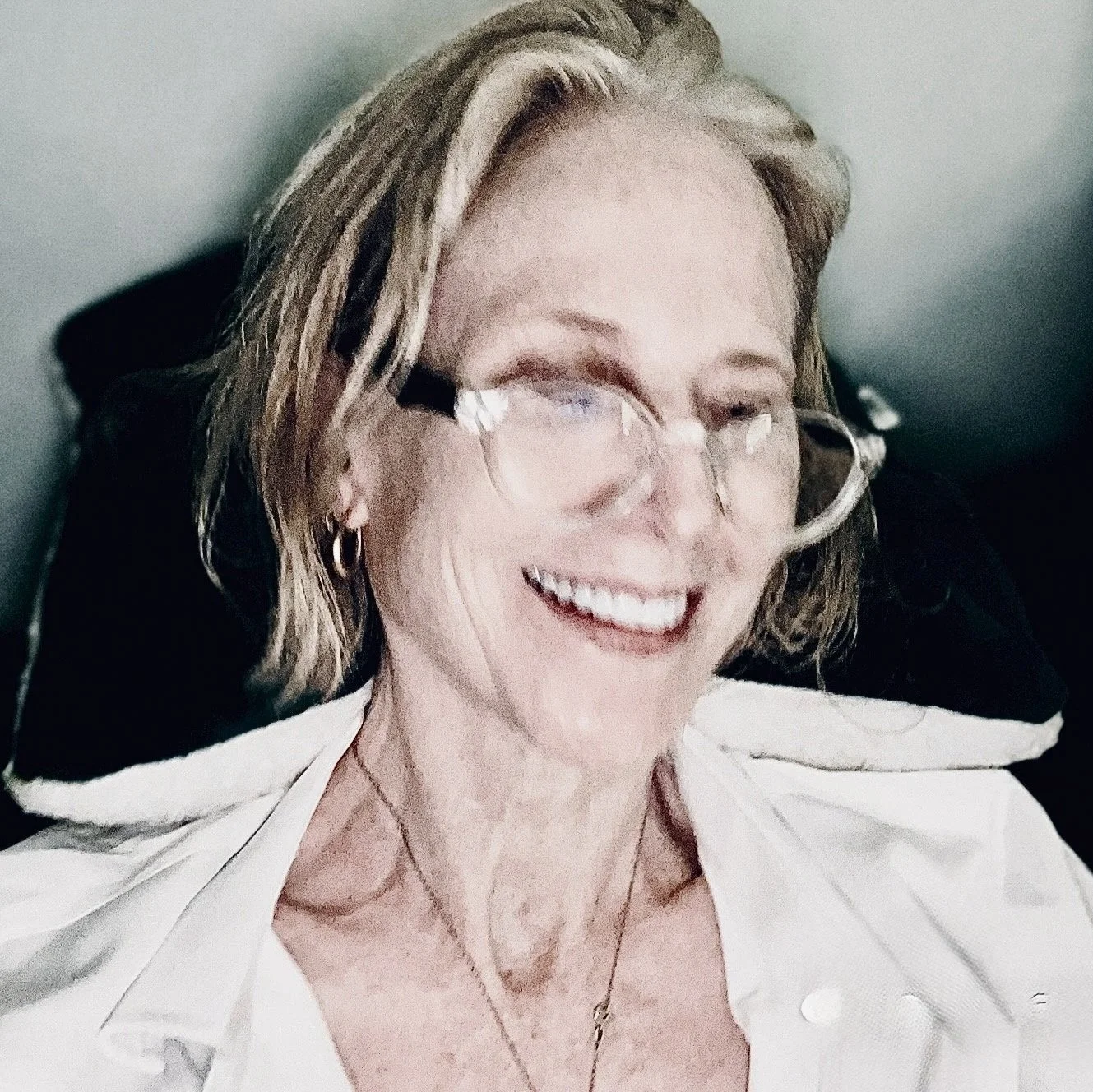 Close-up of a smiling woman wearing glasses, a white shirt, and hoop earrings, with shoulder-length blonde hair, lying back on a black surface.