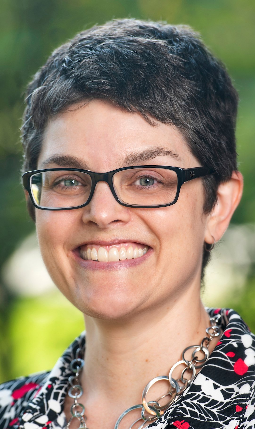 Close-up of woman with short dark hair, black glasses, smiling outdoors in natural light, wearing a patterned top and a chunky silver necklace.