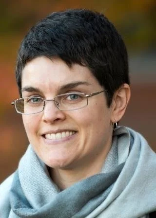 Close-up of a smiling person with short dark hair, wearing glasses and a light-colored scarf outdoors.