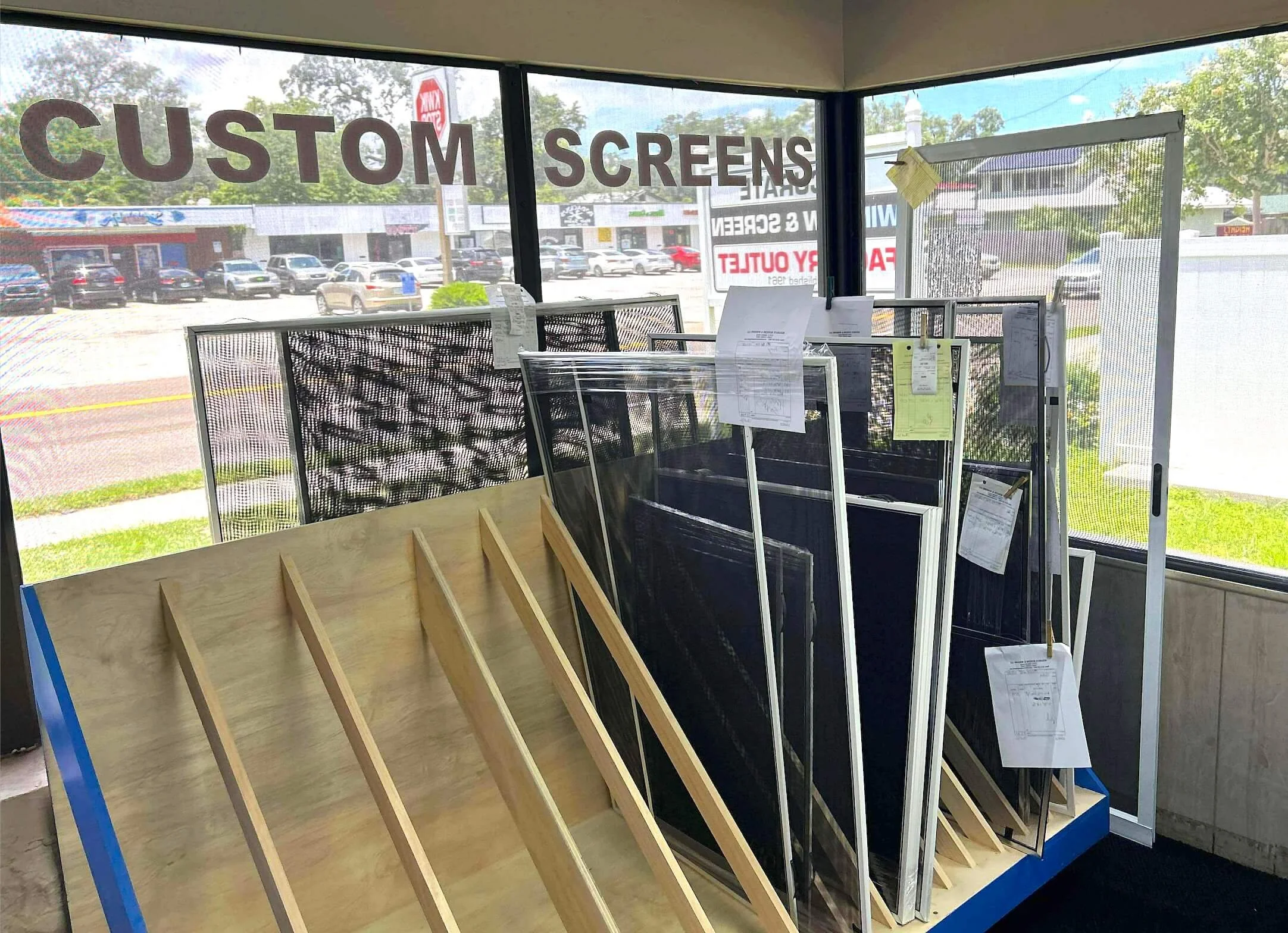 Interior of a screen shop displaying custom-built window screens in various sizes, stacked upright in wooden slots near a large window labeled “CUSTOM SCREENS.”