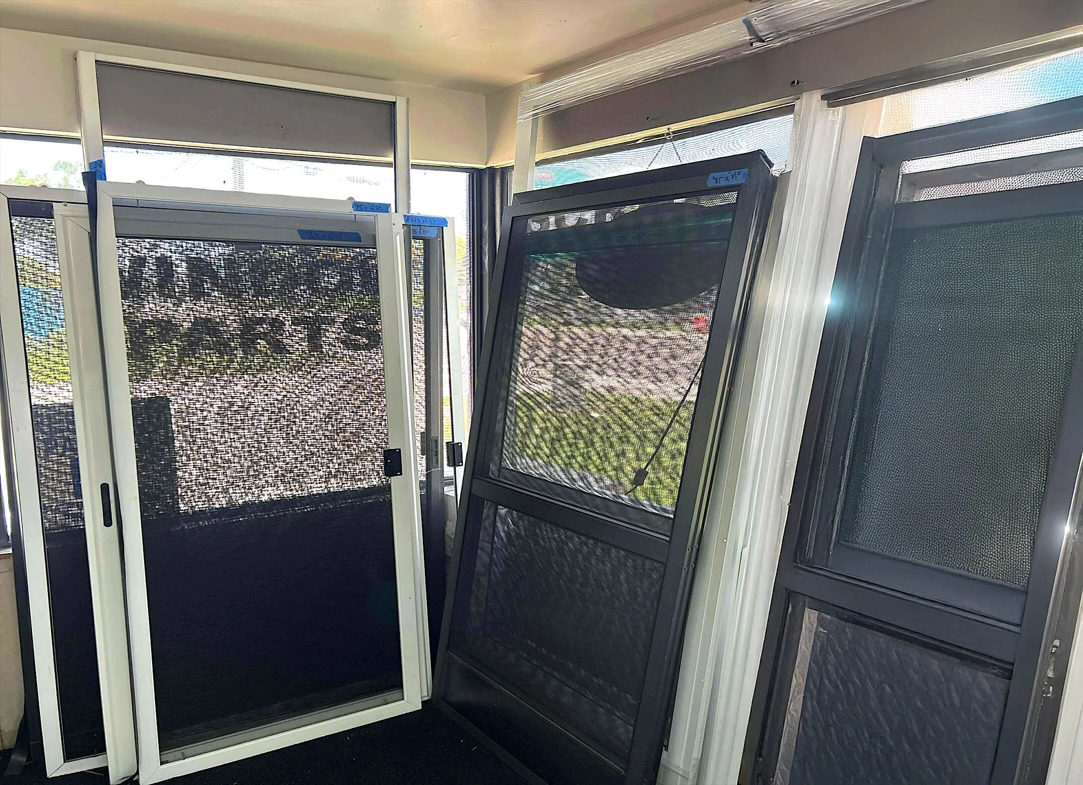 Custom patio and screen doors in various sizes and colors displayed upright inside a showroom, with natural light coming through surrounding windows.