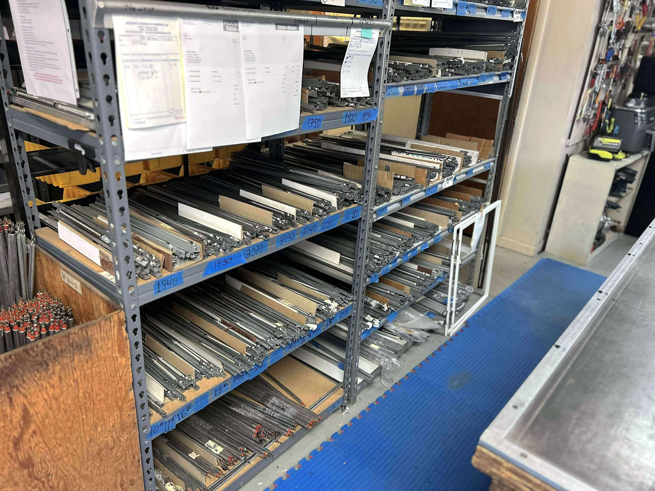 Industrial shelving unit filled with organized window and door hardware parts, including metal tracks and brackets, inside a repair shop with tools and paperwork visible in the background.