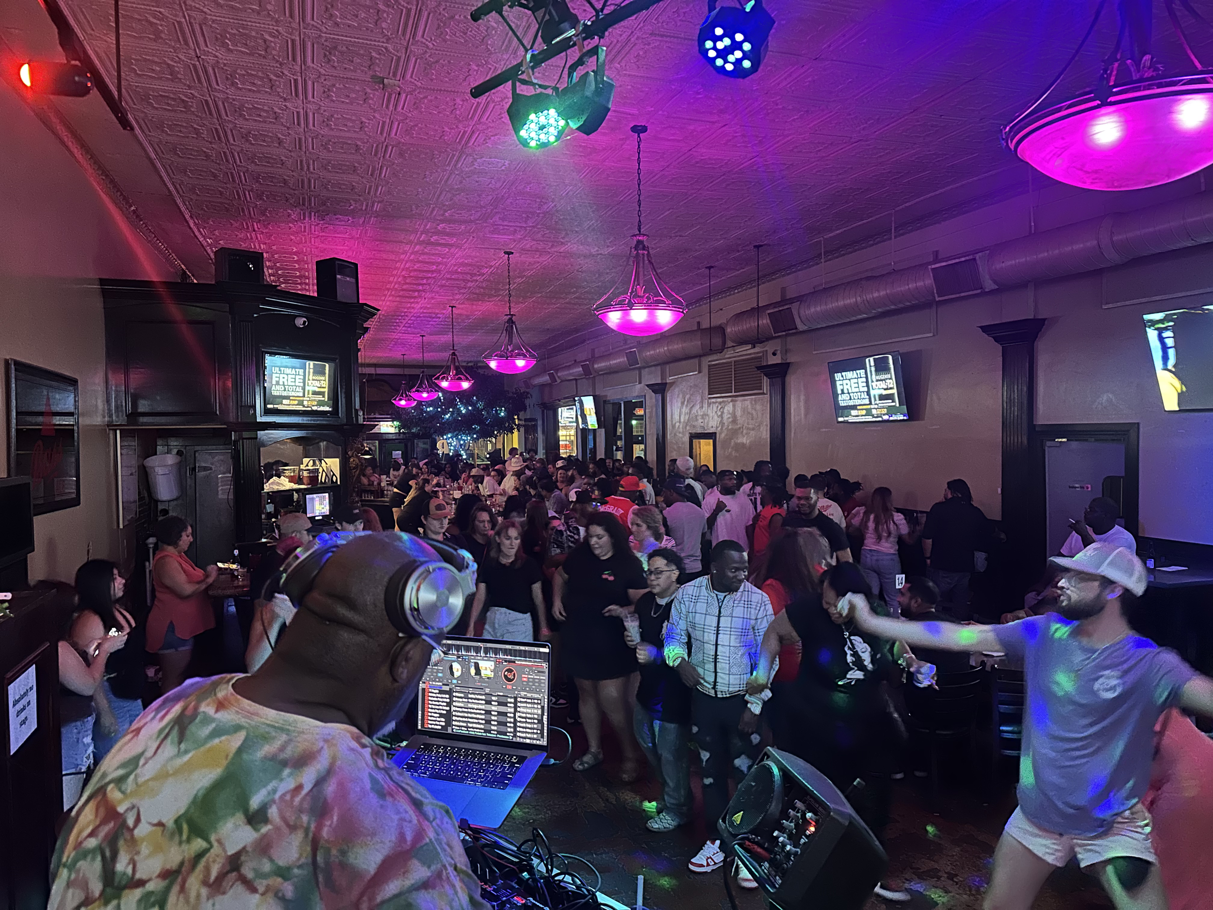 Crowd of people dancing and socializing in a dimly lit nightclub with colorful lighting and a DJ playing music in the foreground.