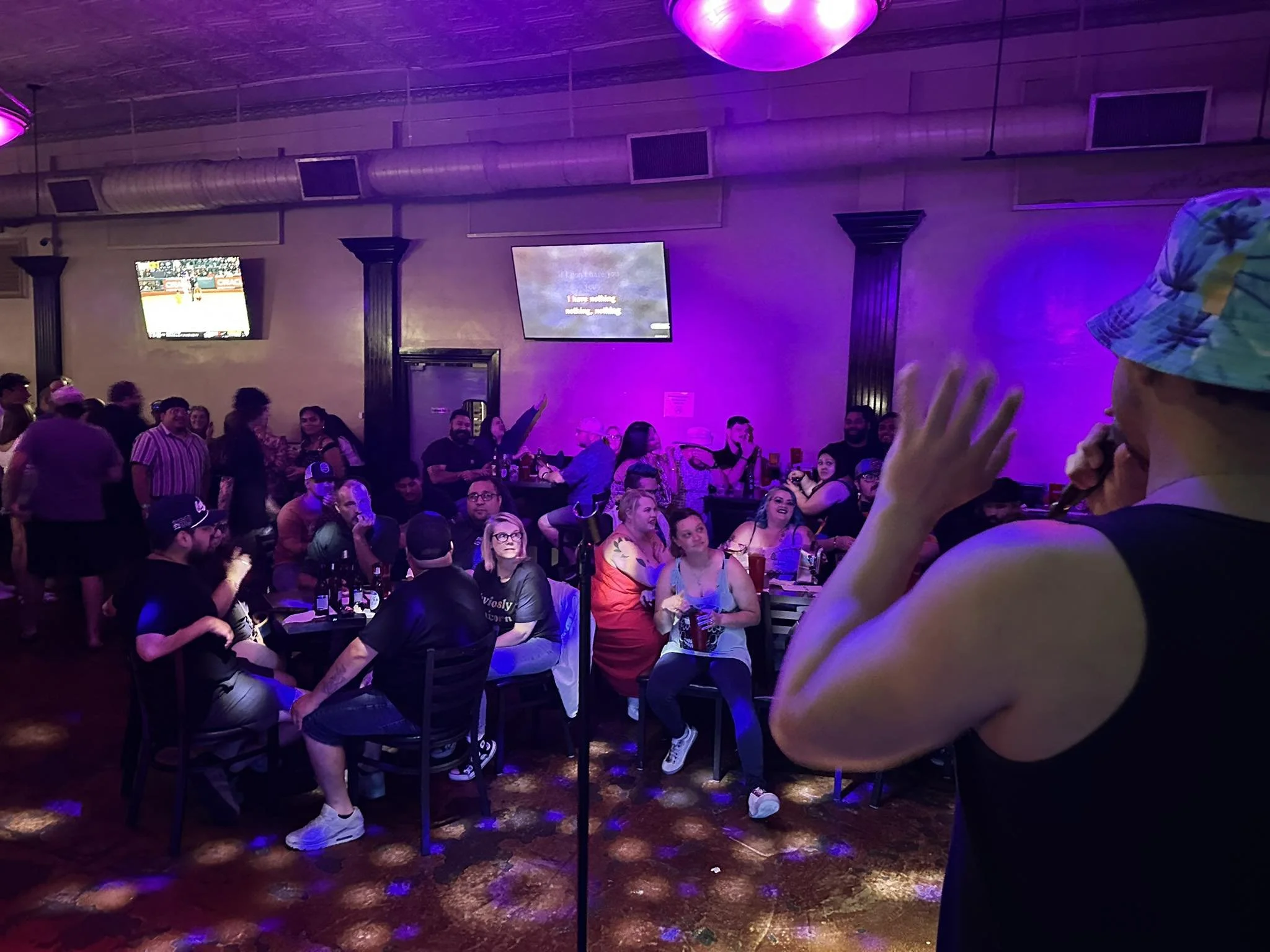 A woman wearing a colorful bucket hat and a sleeveless black top speaks into a microphone, performing in front of an audience in a dimly lit bar or club with purple lighting. The audience is sitting and standing, watching and enjoying the performance.