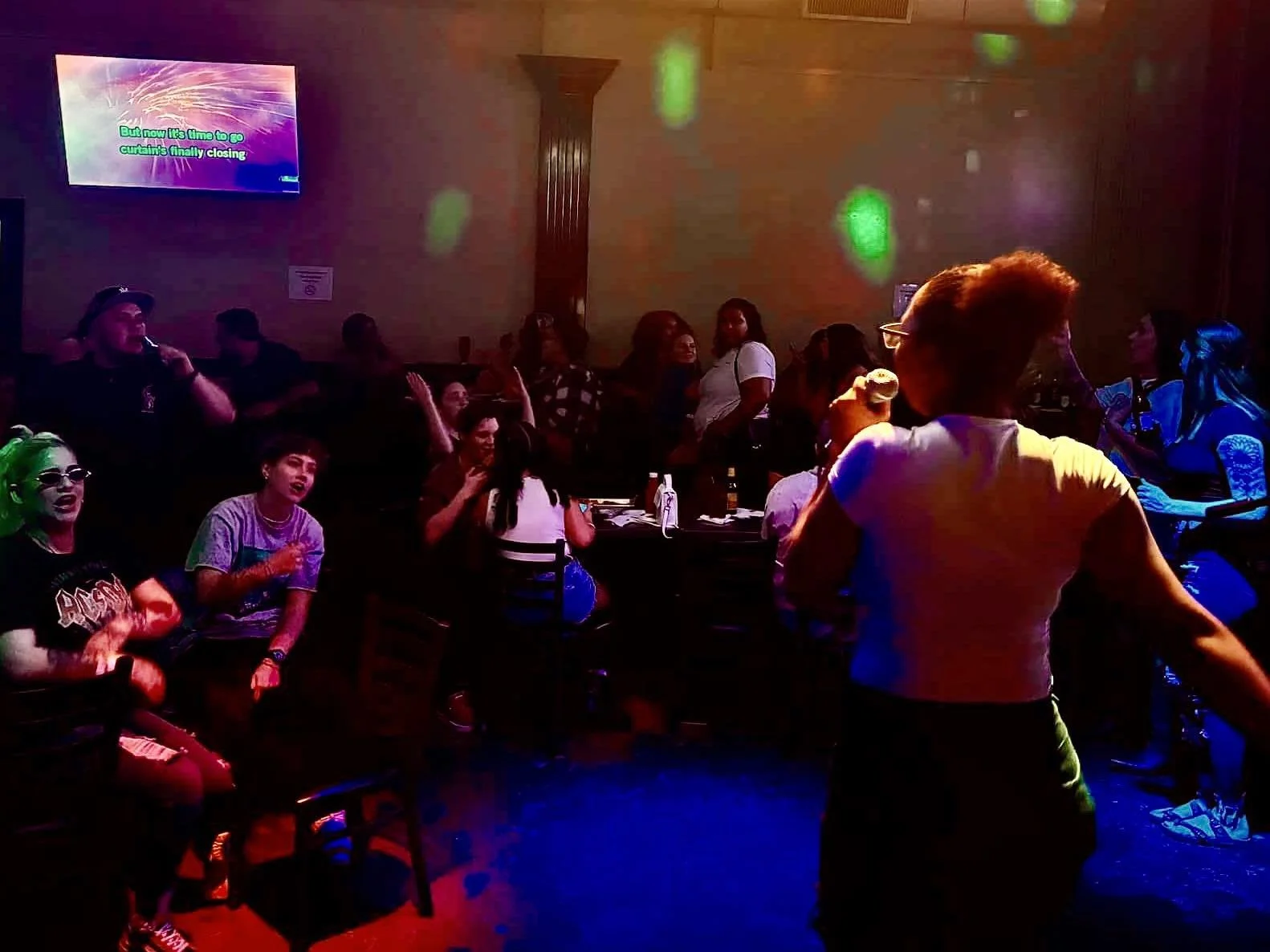 People singing karaoke in a dimly lit bar with colorful lights and a video screen displaying song lyrics.