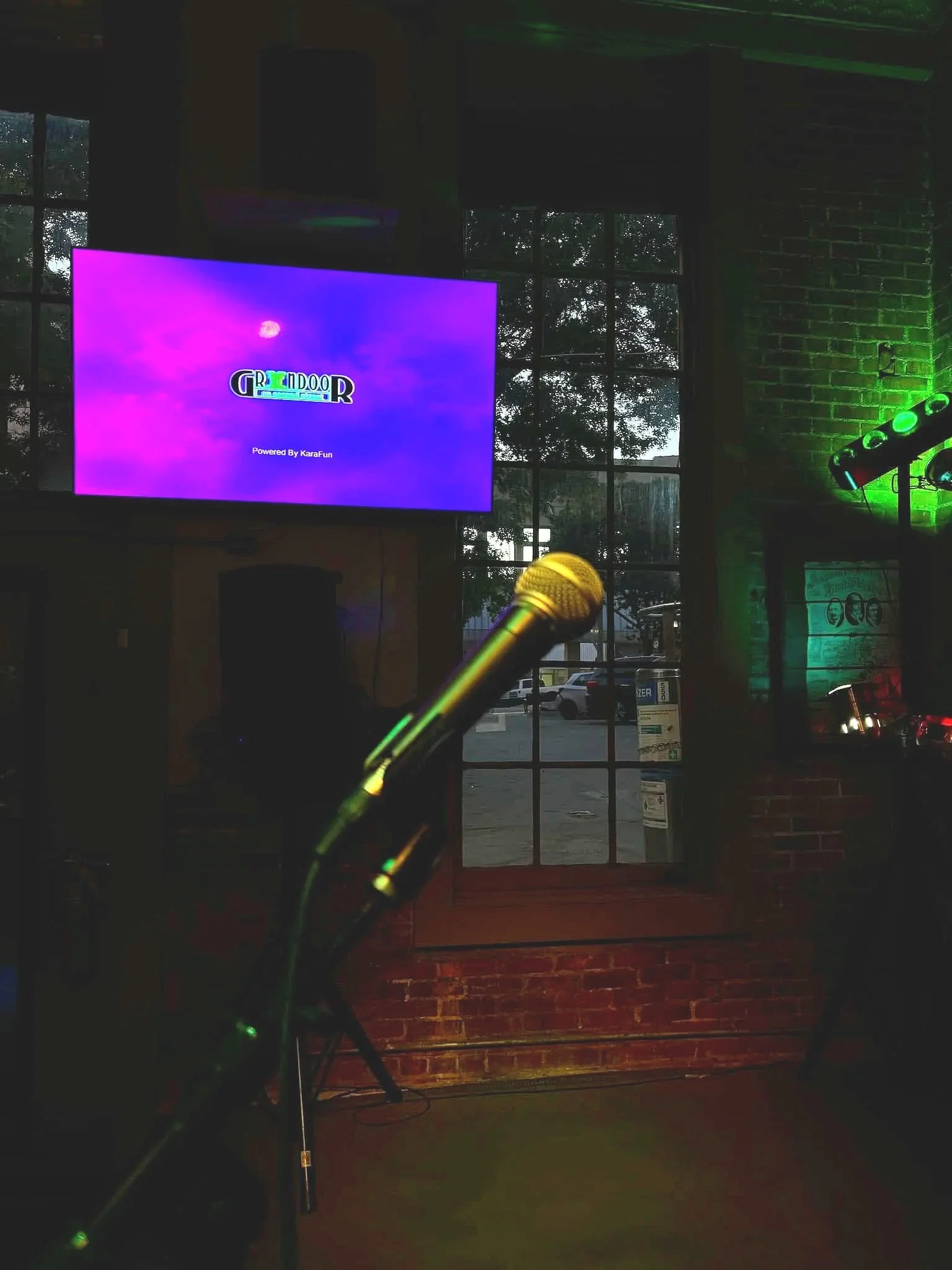Microphone in front of a large screen displaying colorful graphics, with a brick wall, windows showing an outdoor parking lot, and green lighting in the background, indicating a performance or karaoke venue.