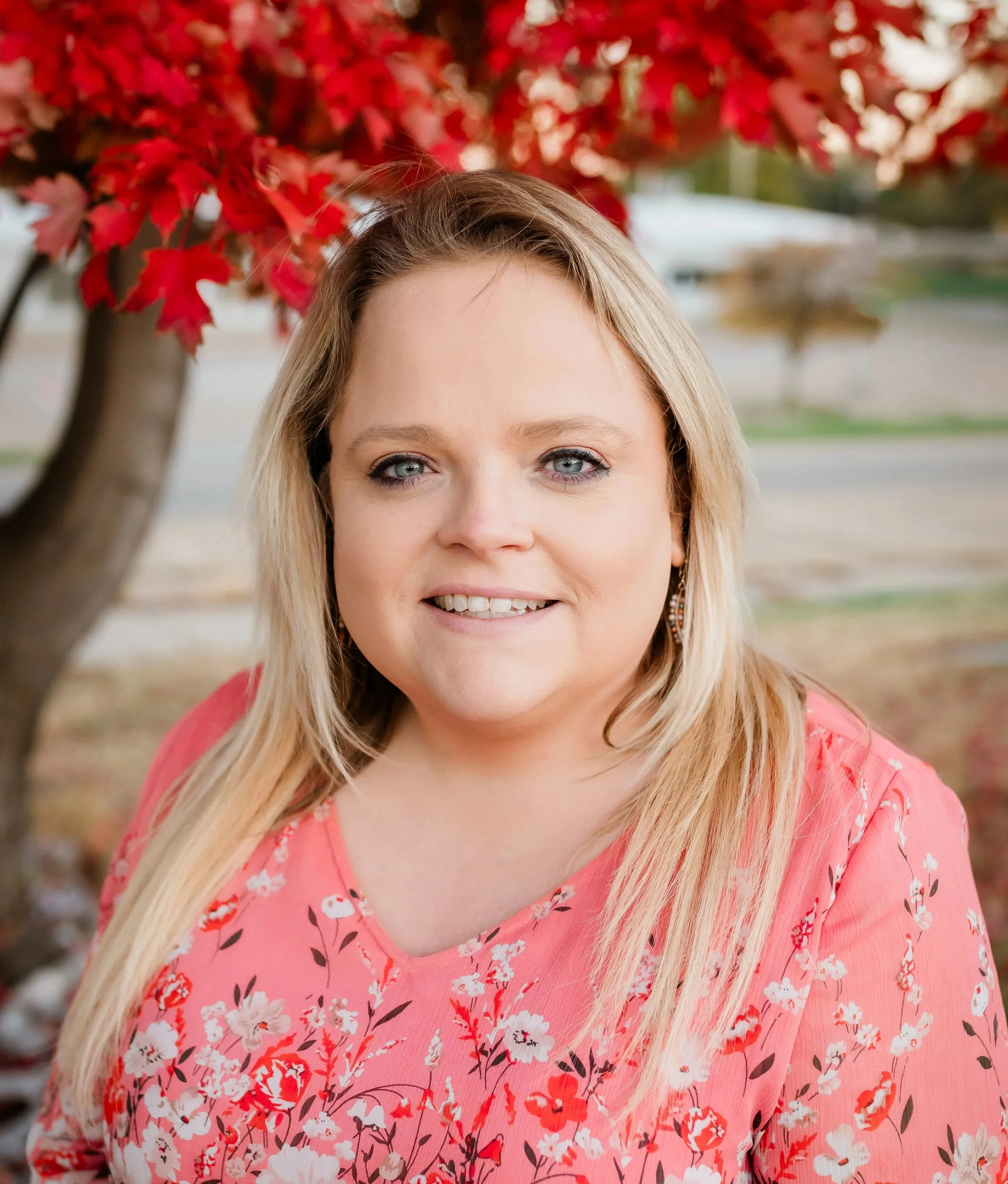 A woman with blonde hair, blue eyes, and light skin, wearing a pink floral blouse, standing outdoors in front of a tree with red leaves.