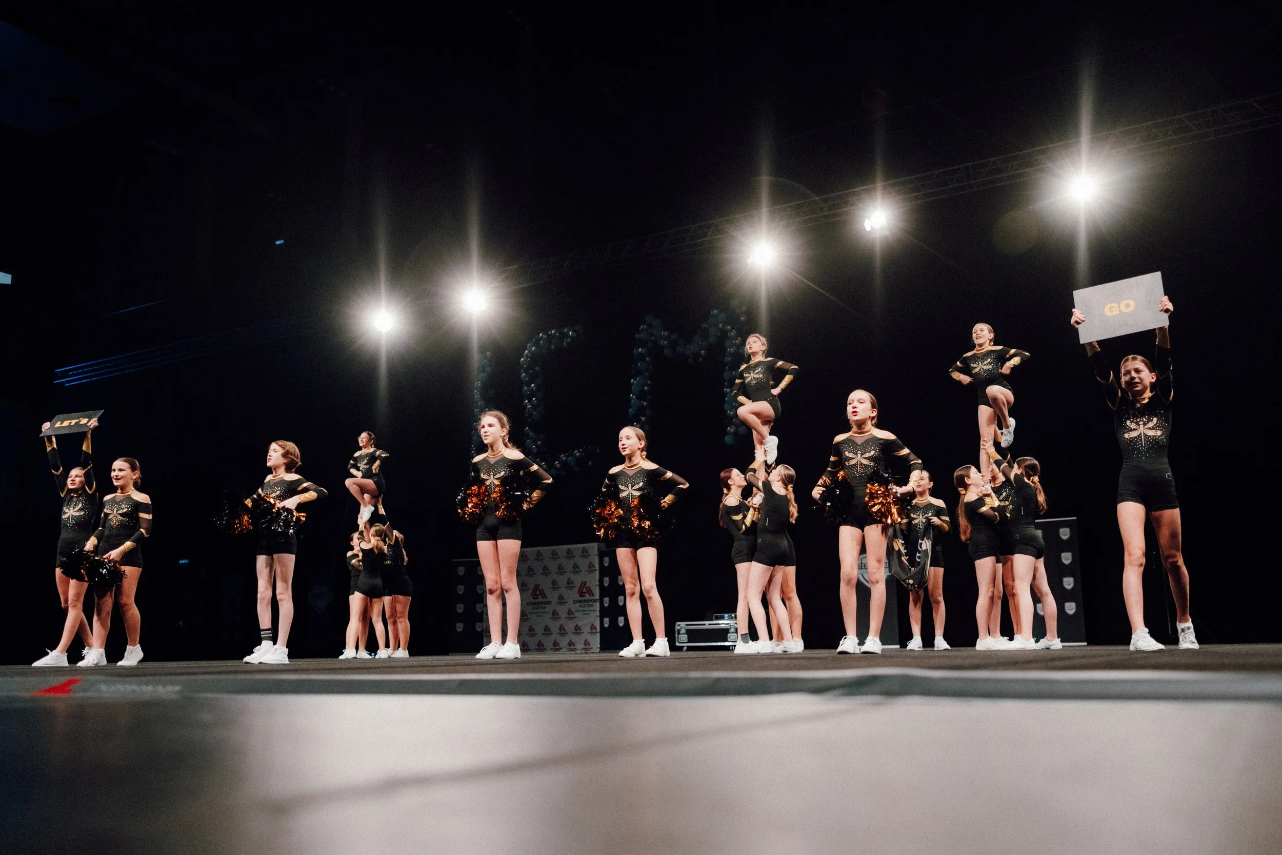 Jugendliche Cheerleader bei einer Wettkampfvorführung auf der Bühne, einige halten Schilder mit Aufschriften, andere posieren oder führen Pyramiden aus, im Hintergrund sind Ballons und Bühnenbeleuchtung.