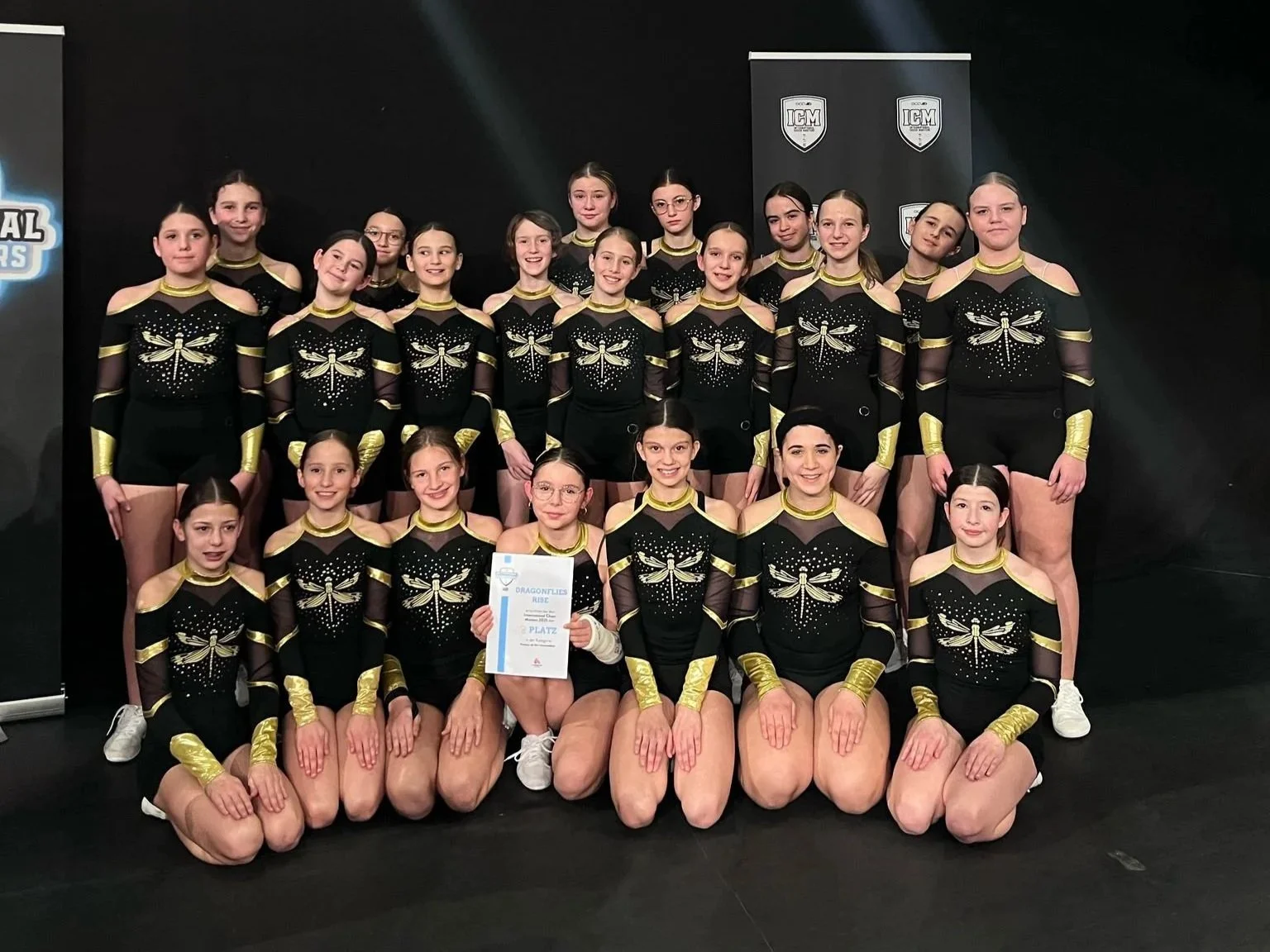 Eine Gruppe junger Mädchen in schwarzen Cheer-Outfits mit goldenen Akzenten und Libellen-Design, die auf einer Bühne sitzen und eine Urkunde halten, vermutlich nach einem Wettbewerb, in einer Turnhalle oder auf einer Bühne bei einer Auszeichnung.