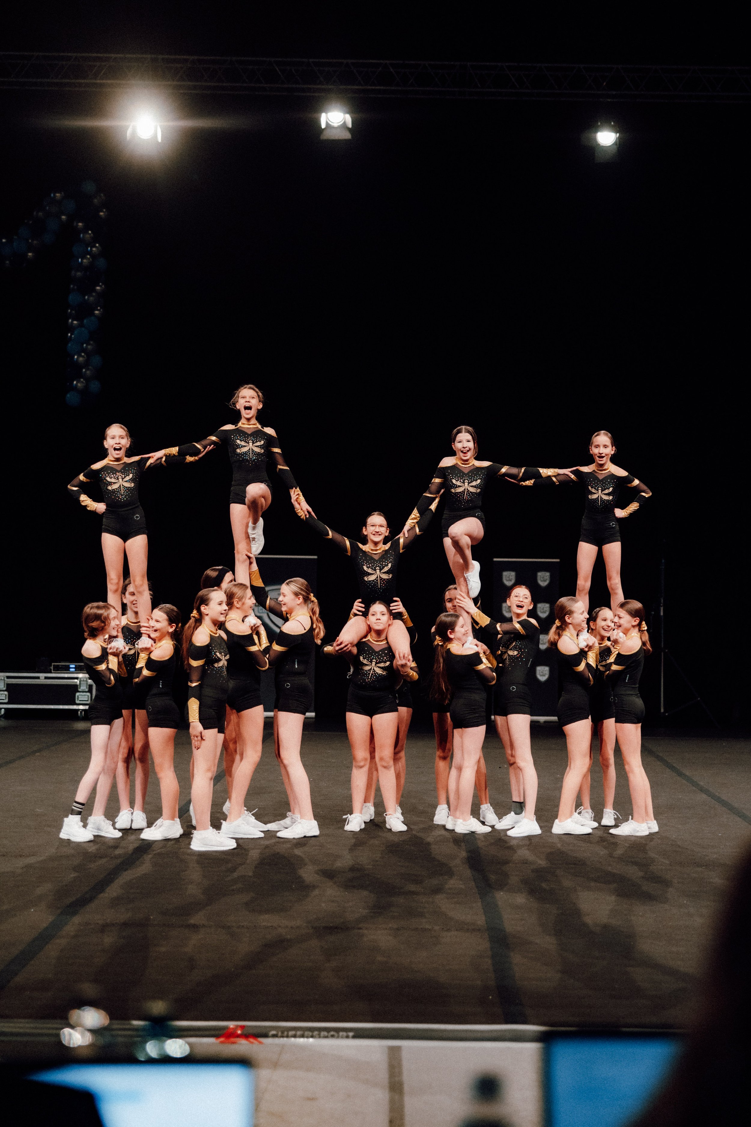 Jugendliche Cheerleader zeigen eine akrobatische Formation (Pyramide) auf einer Bühne, umgeben von anderen Cheerleadern in schwarzen und goldfarbenen Uniformen.