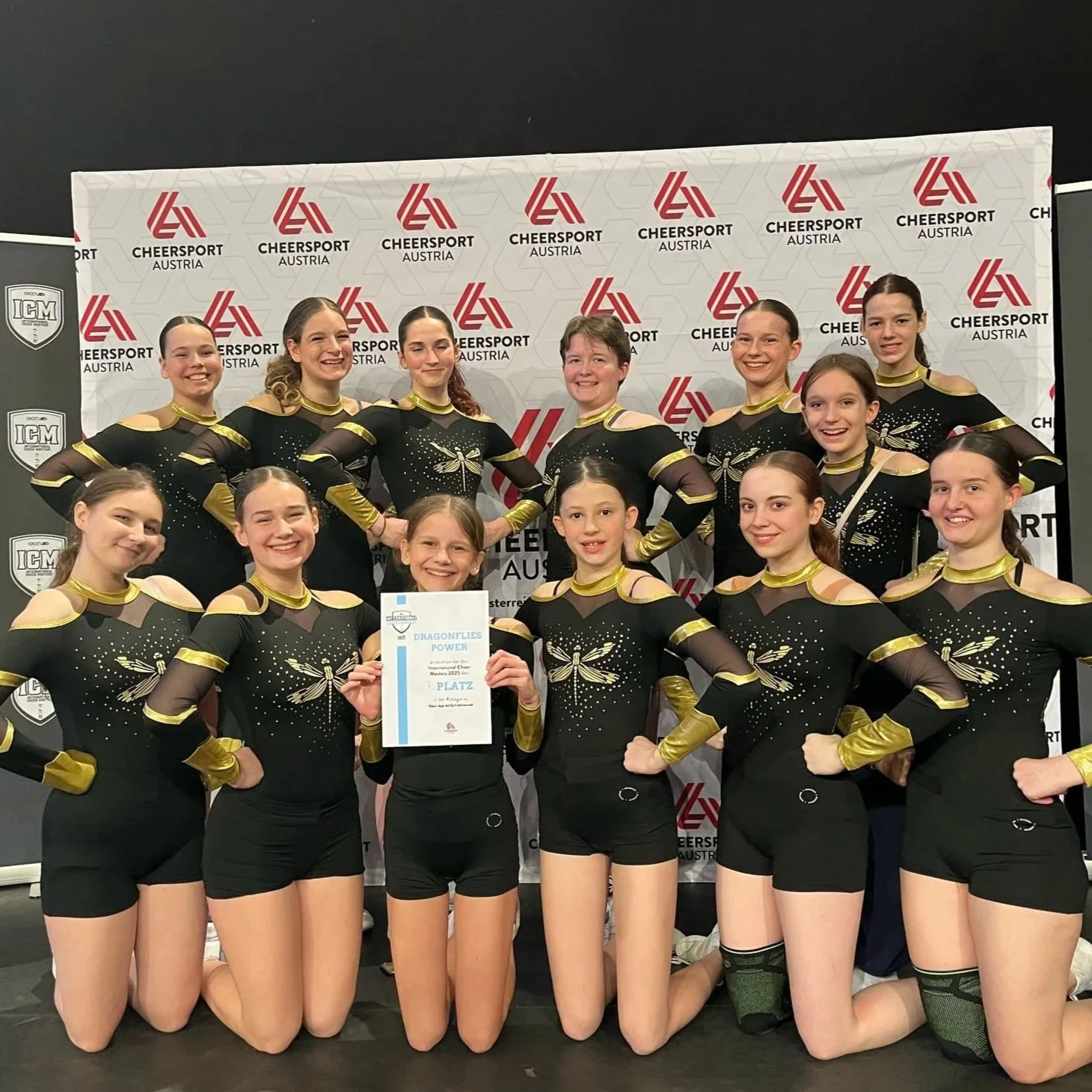 Eine Gruppe von16 jungen Mädchen in schwarzen Turnanzügen mit goldfarbenen Details posiert vor einem Hintergrund mit Cheerleading-Logos. Eines Mädchen hält eine Urkunde mit der Aufschrift 'Dragons Power' und '2. Platz' in den Händen.