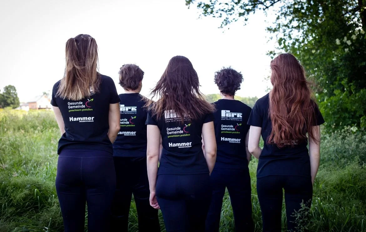 Fünf Frauen stehen mit dem Rücken zueinander in einem grünen Freiraum, alle tragen schwarze T-Shirts mit Logos und Texten.