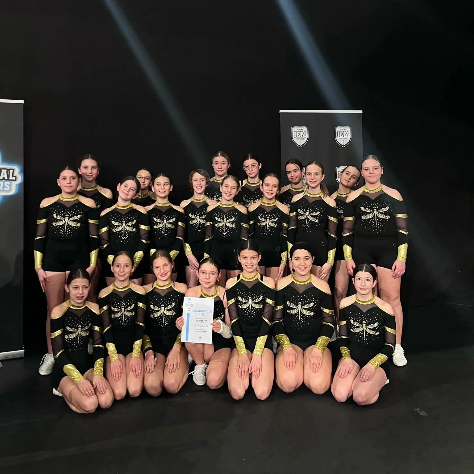Eine Gruppe junger Mädchen in schwarzem und goldenem Turnanzug, die eine Cheerleading-Performance im Wettbewerb gemacht haben und einen Urkunde halten.