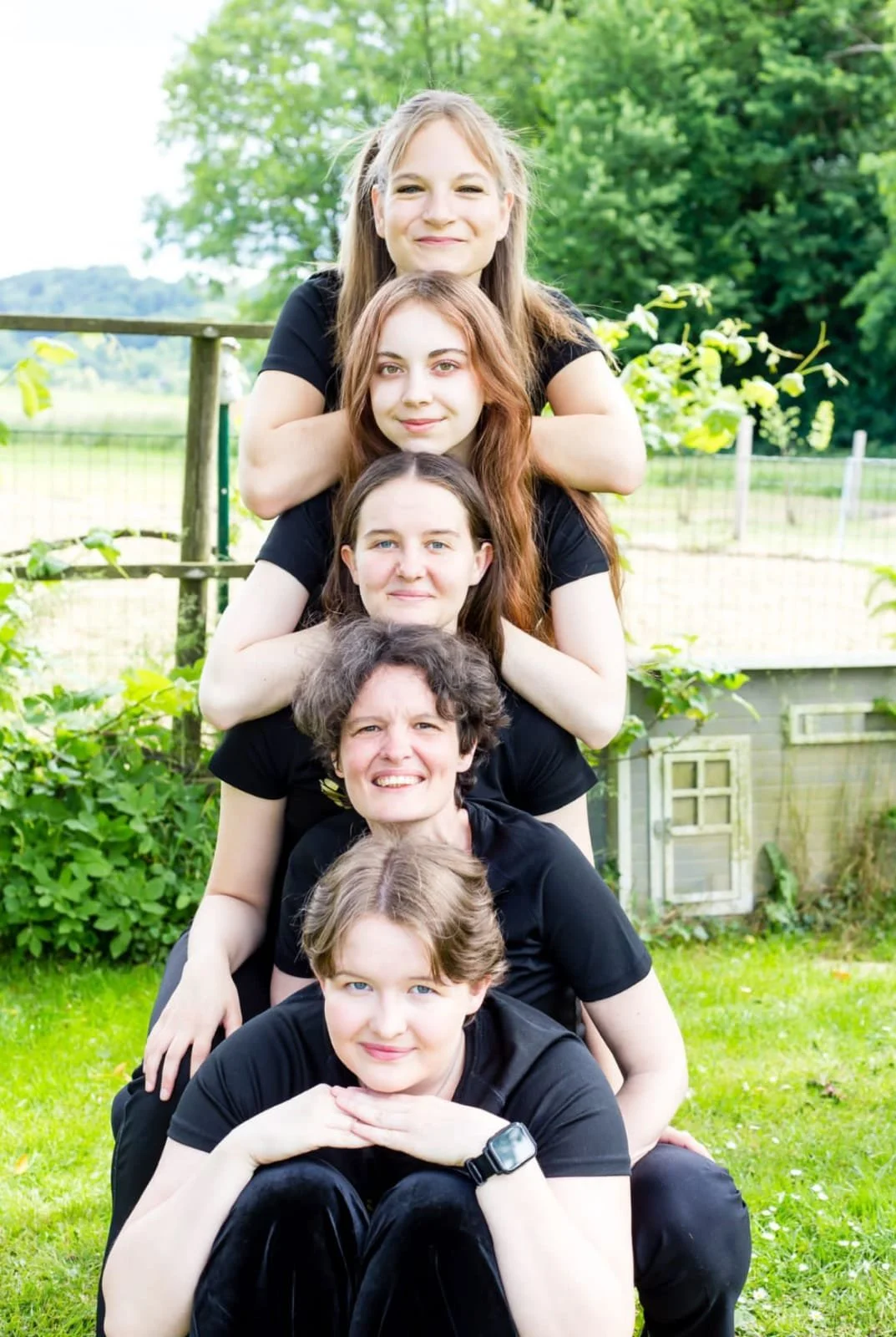 Sechs Frauen sitzen in einer Pyramide im Garten vor einem Baum und einem Holzzaun. Sie tragen alle schwarze T-Shirts, und es ist eine grüne, ländliche Umgebung sichtbar.