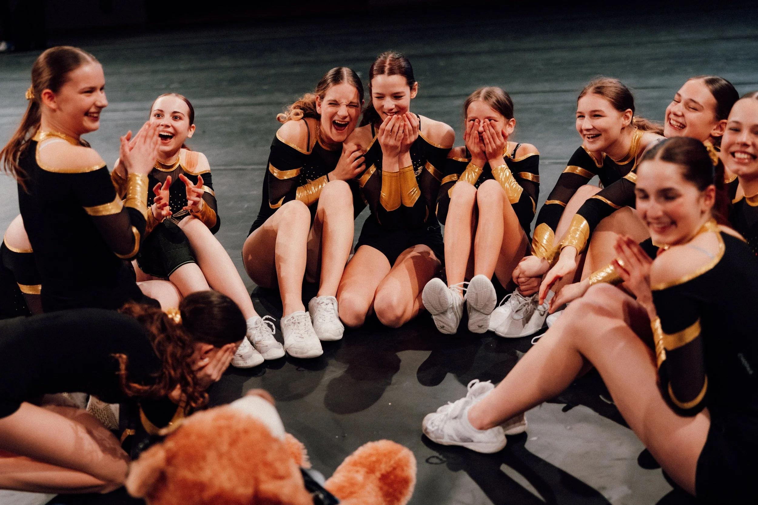 Junge Cheerleaderinnen, die auf der Bühne sitzen und lachen, in schwarzen und goldenen Uniformen, eine freut sich besonders, umgeben von ihren Teamkameraden.