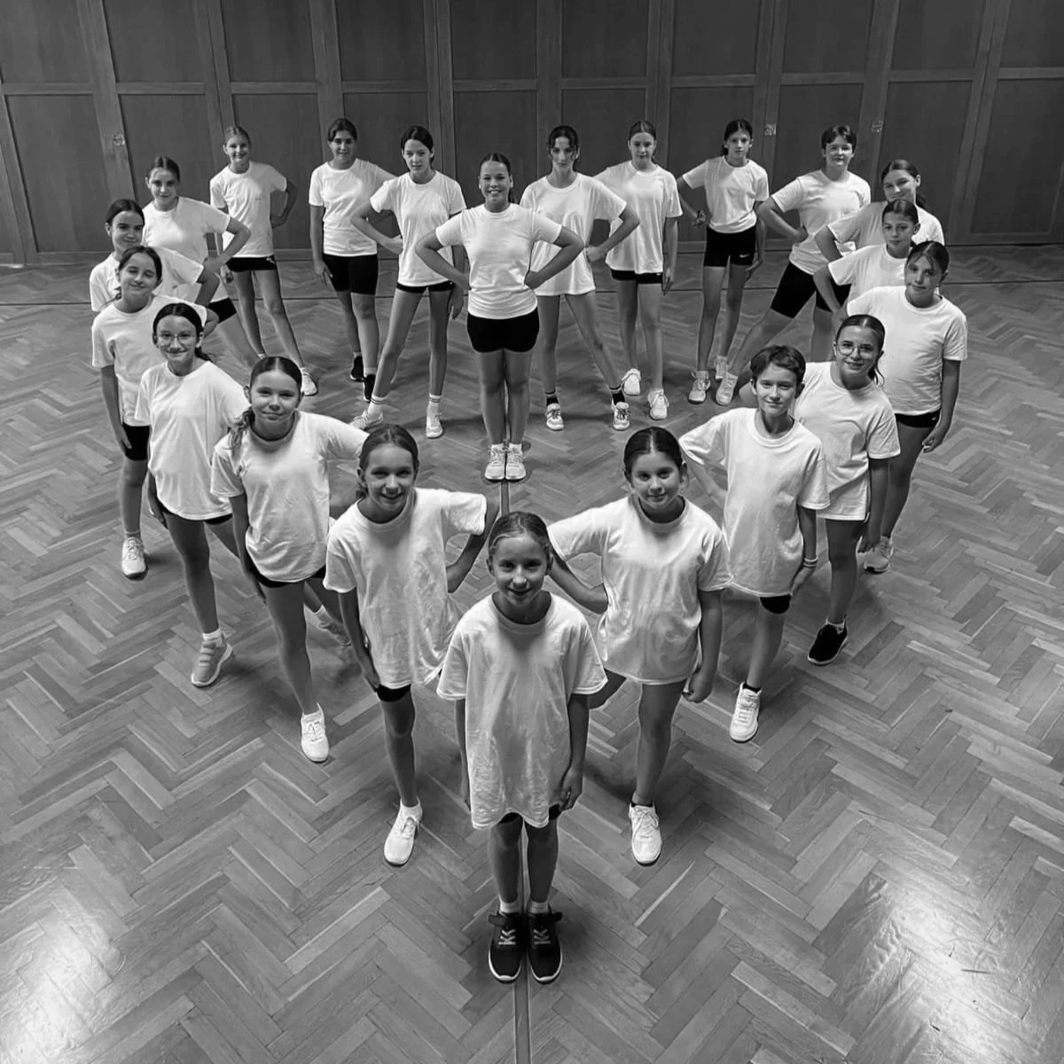 Eine Gruppe junger Mädchen in weißen T-Shirts und Shorts, die in einer Sporthalle in Herzform aufgestellt sind.