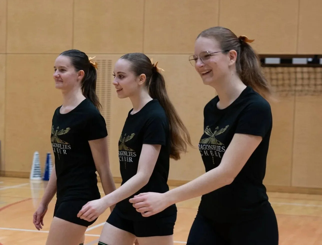 Drei junge Frauen in schwarzen Sportshirts und Shorts, die in einer Turnhalle lächeln und sich unterhalten.