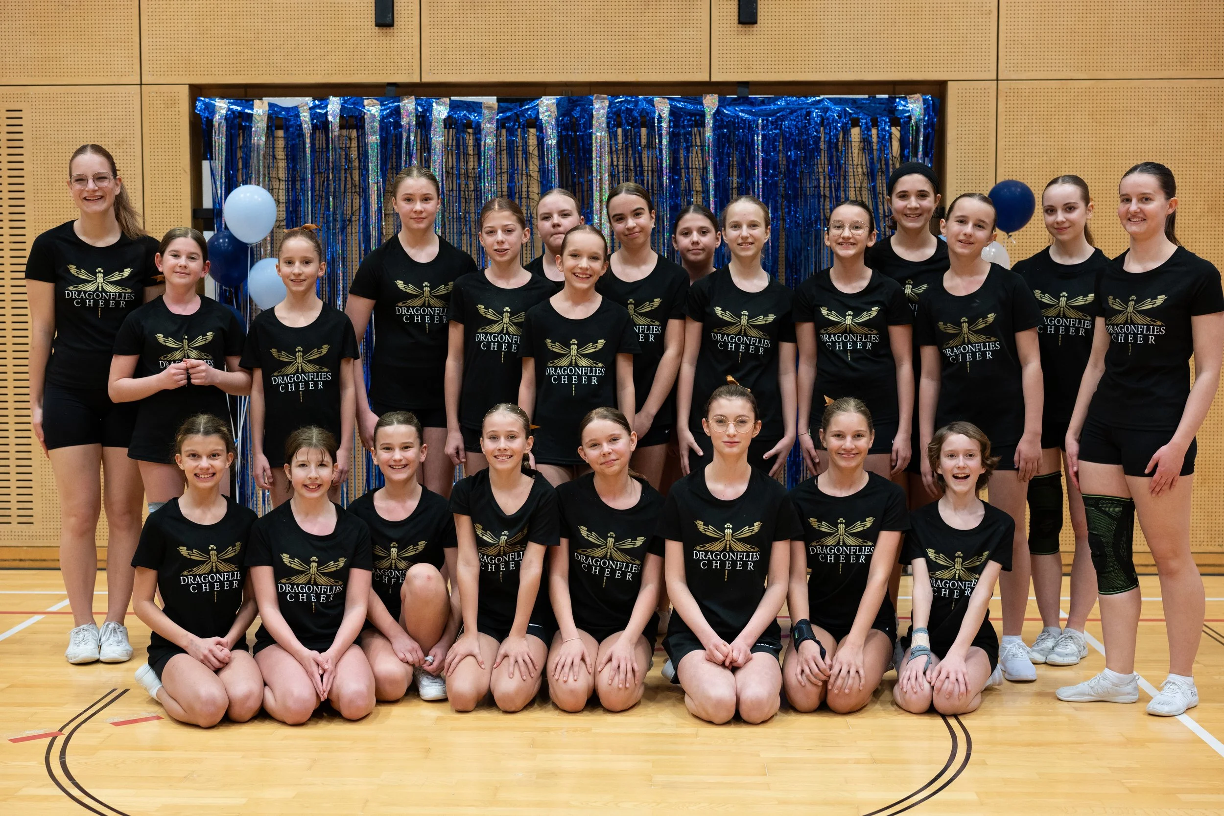 Gruppe junger Mädchen in schwarzen Cheerleading-Uniformen mit dem Text "Dragonflies Cheer", posieren in einer Turnhalle vor einem blauen und silbernen Hintergrund.